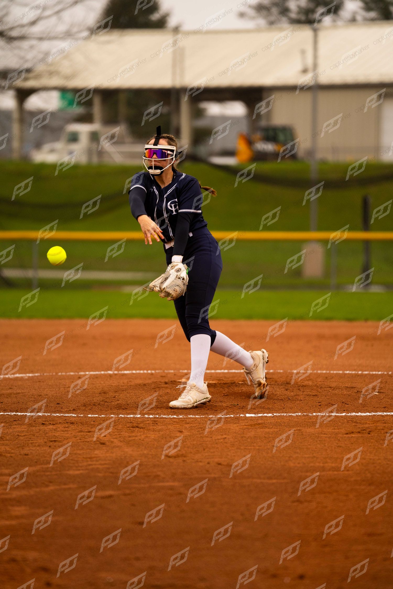 Female softball pitcher in navy uniform delivering a yellow ball in windmill motion on clay field.