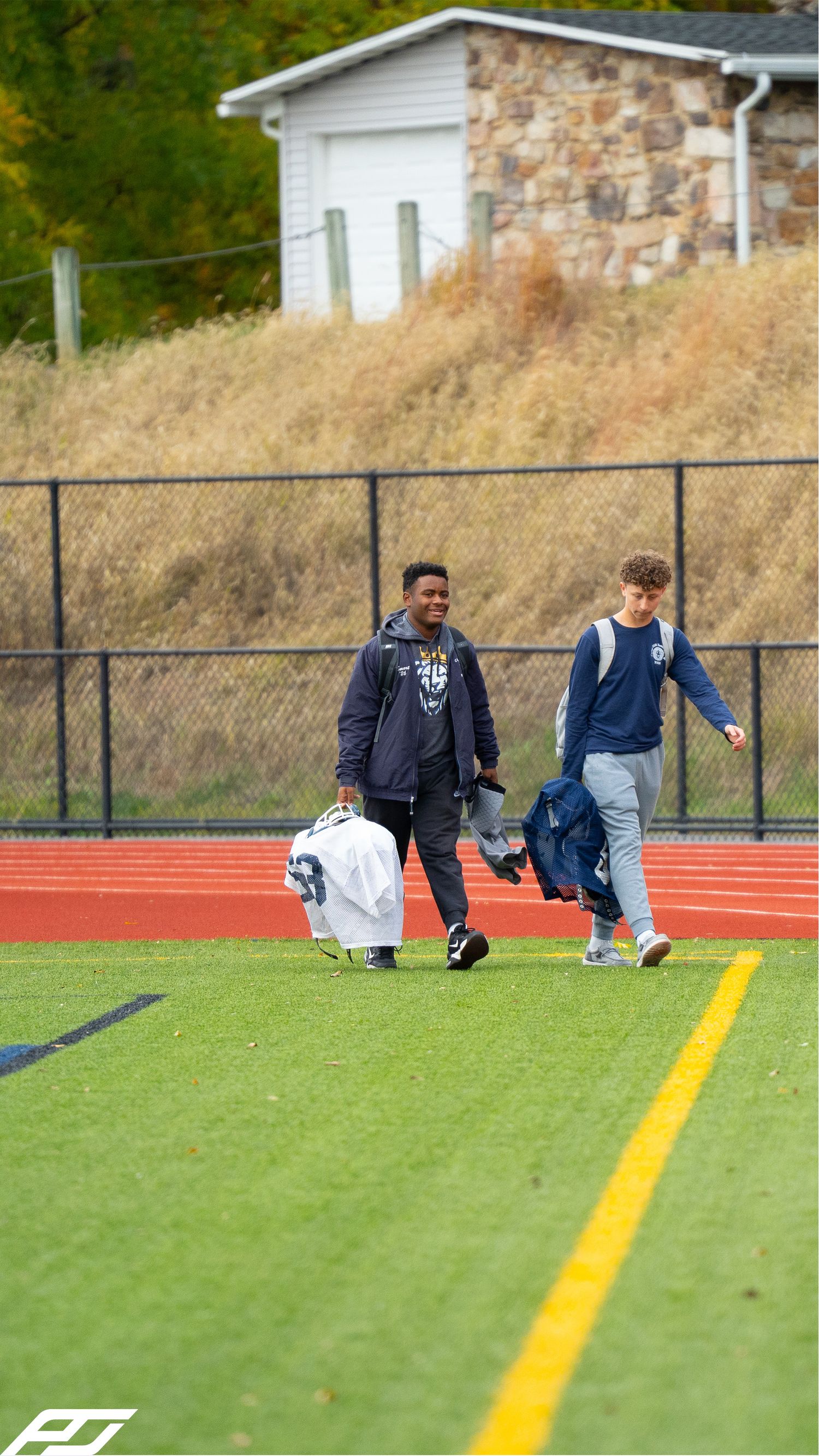 Two youth football players in navy uniforms carry gear across a green turf field during an autumn game day.