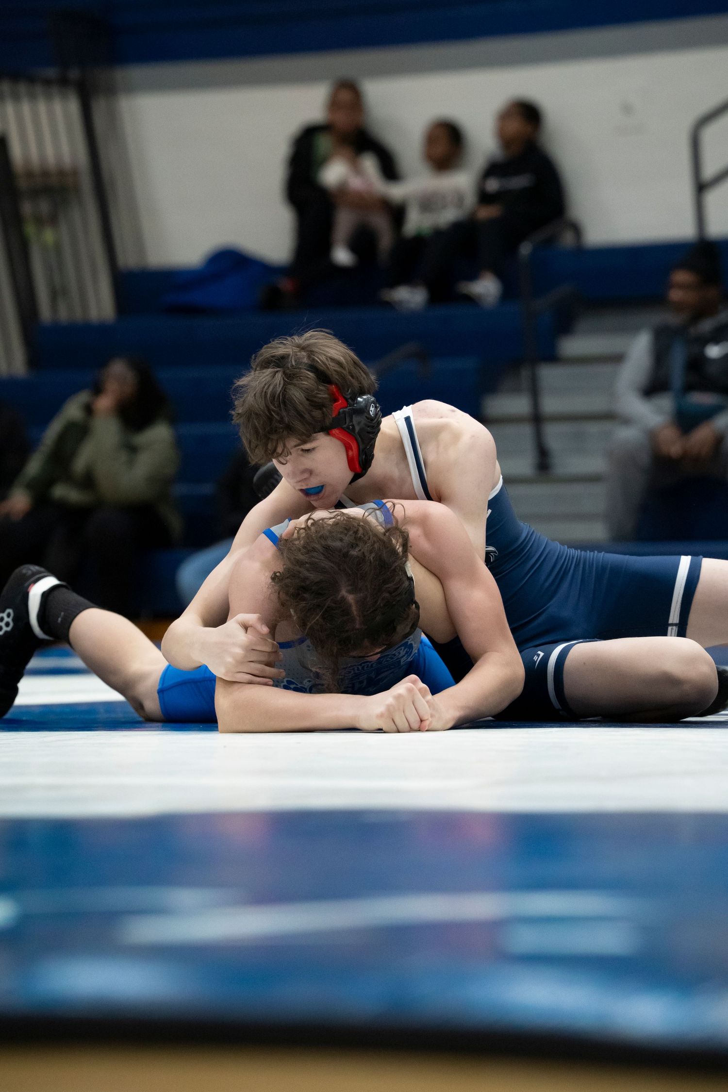 Young wrestler in red headgear pins opponent to the mat during a competitive indoor wrestling match.
