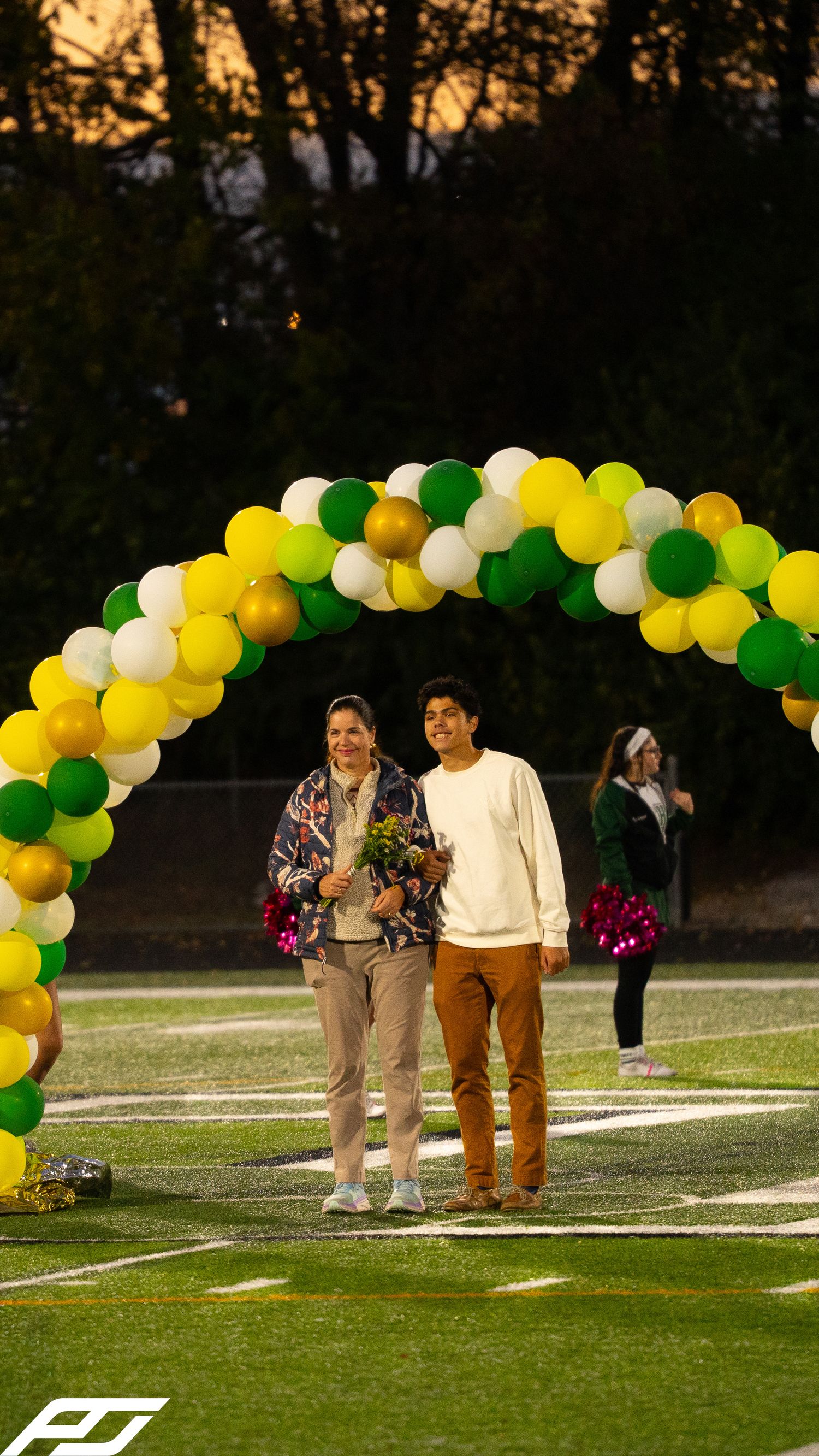 Senior student walks with parent under green and yellow balloon arch during evening field recognition ceremony.