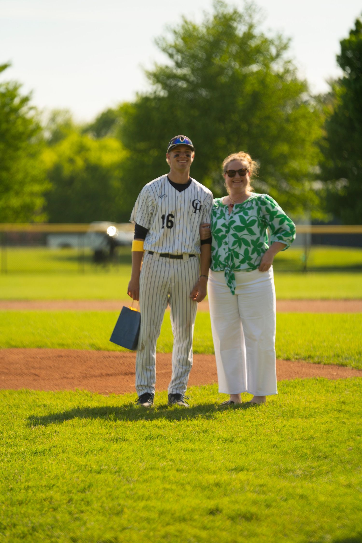 Baseball player #16 in pinstripe uniform stands with a woman on a green baseball field on a sunny day.