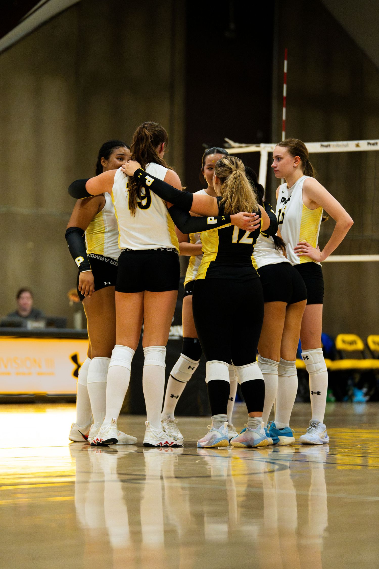 College volleyball players in black and gold uniforms huddle together on the court during a competitive match.