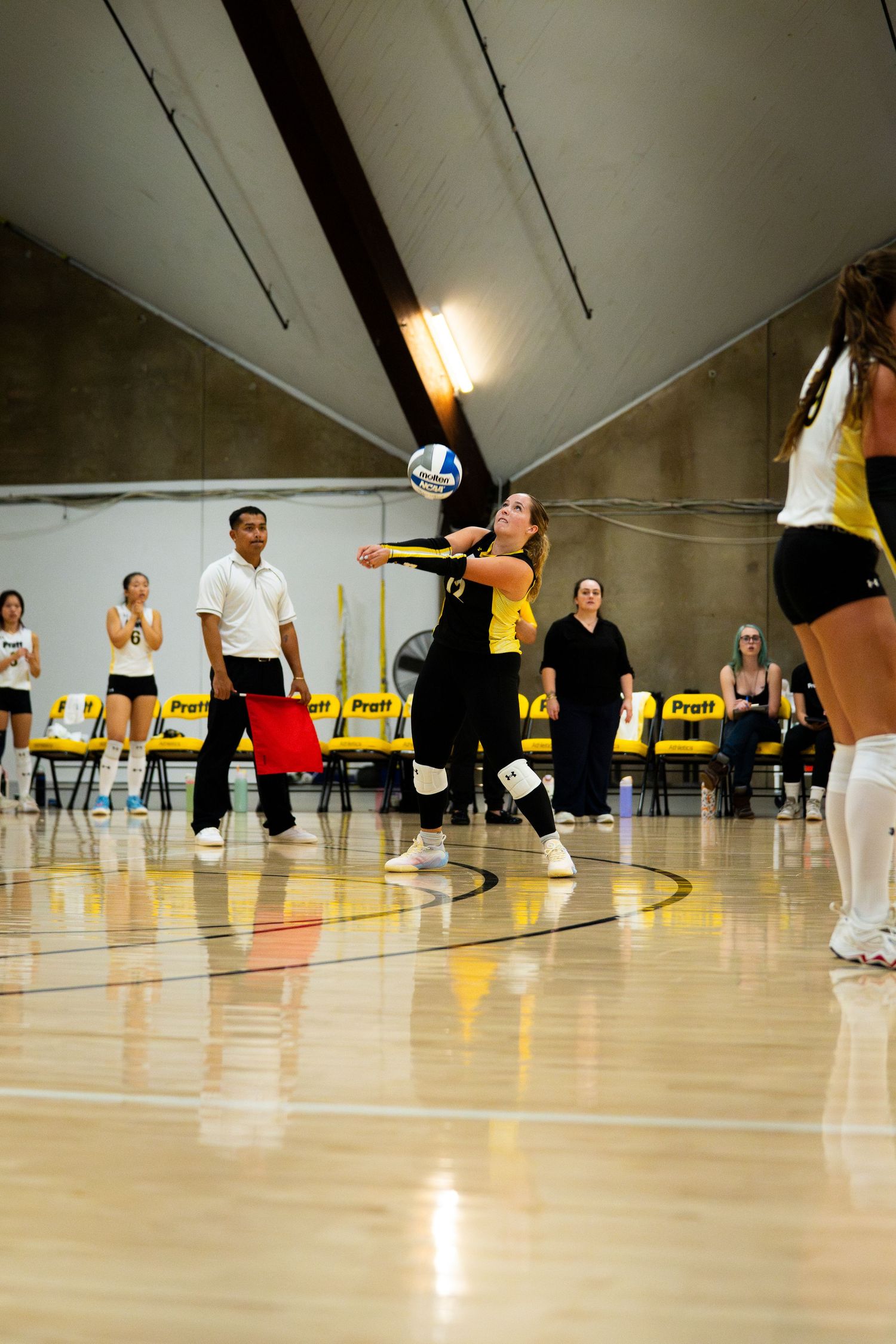 Female volleyball player performs a dig during an indoor match, wearing black and gold uniform on a hardwood court.