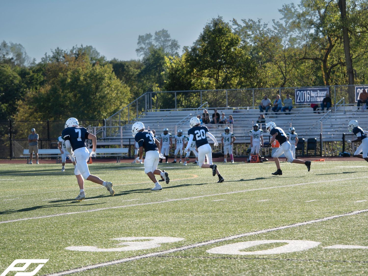 Youth football players in navy uniforms run downfield during a sunny outdoor game on turf.
