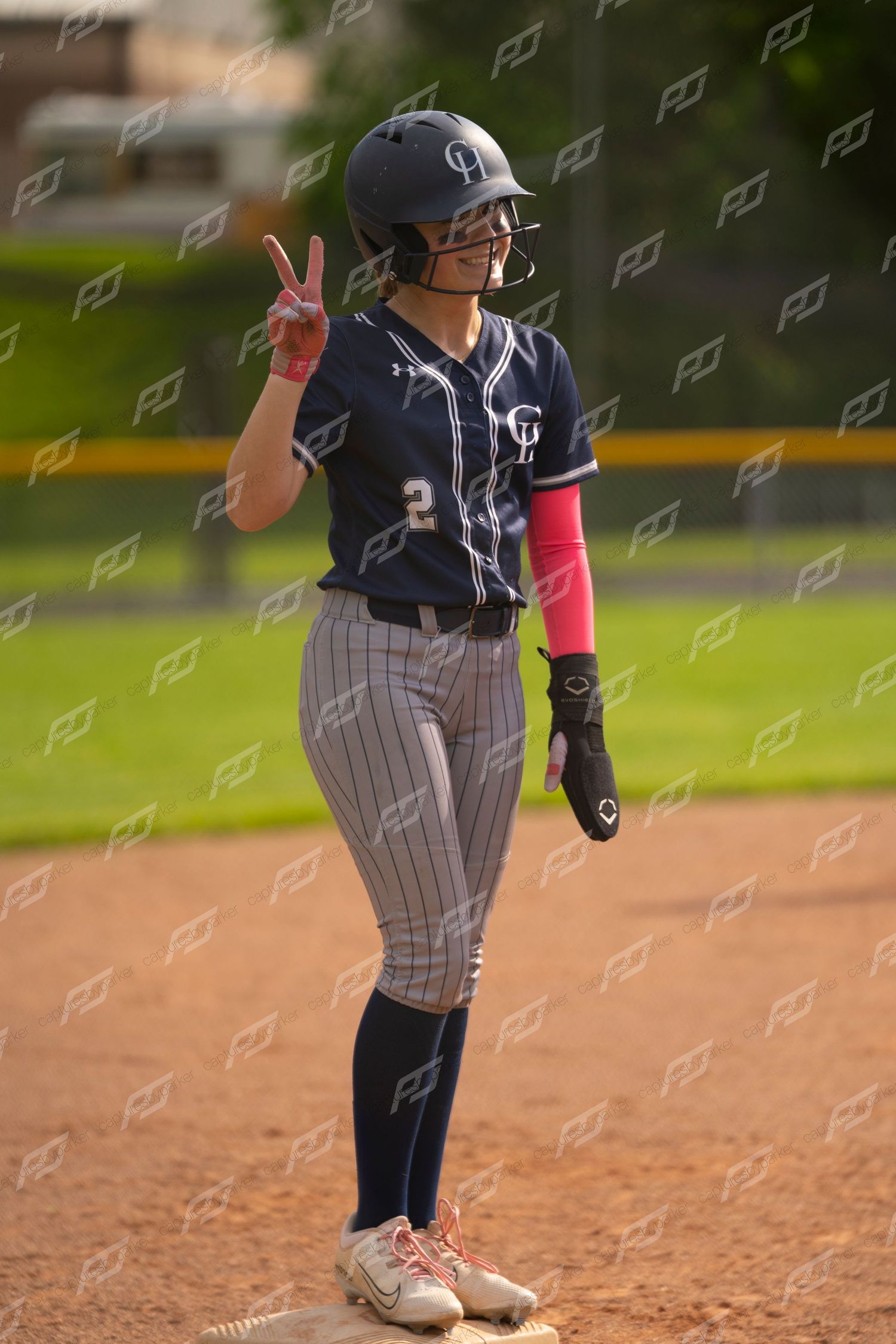 Female softball player in navy uniform holds up a peace sign while standing on second base.