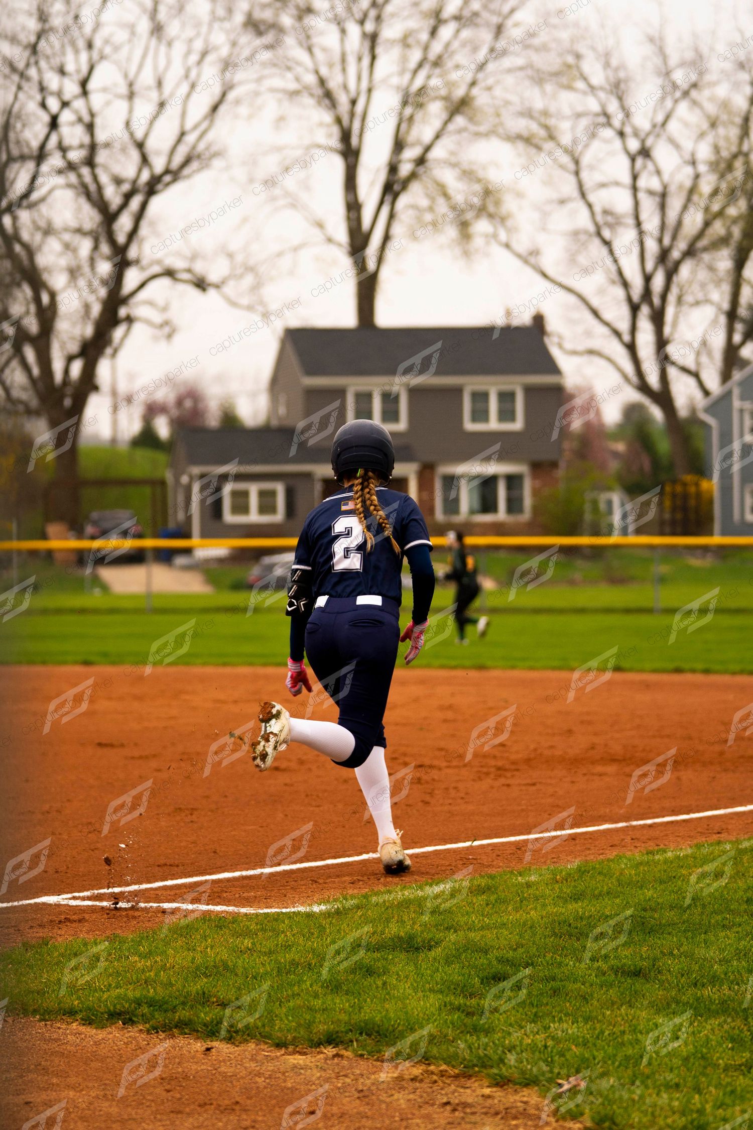 Softball player number 2 in navy uniform runs toward first base on clay infield during a game.