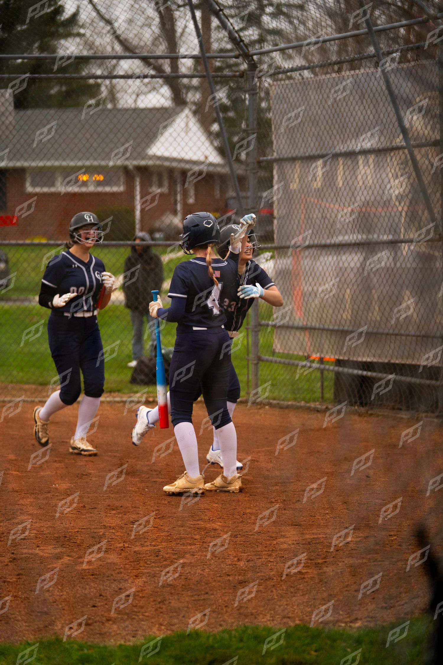 Female softball players in navy uniforms celebrate together near dugout fence after scoring run.