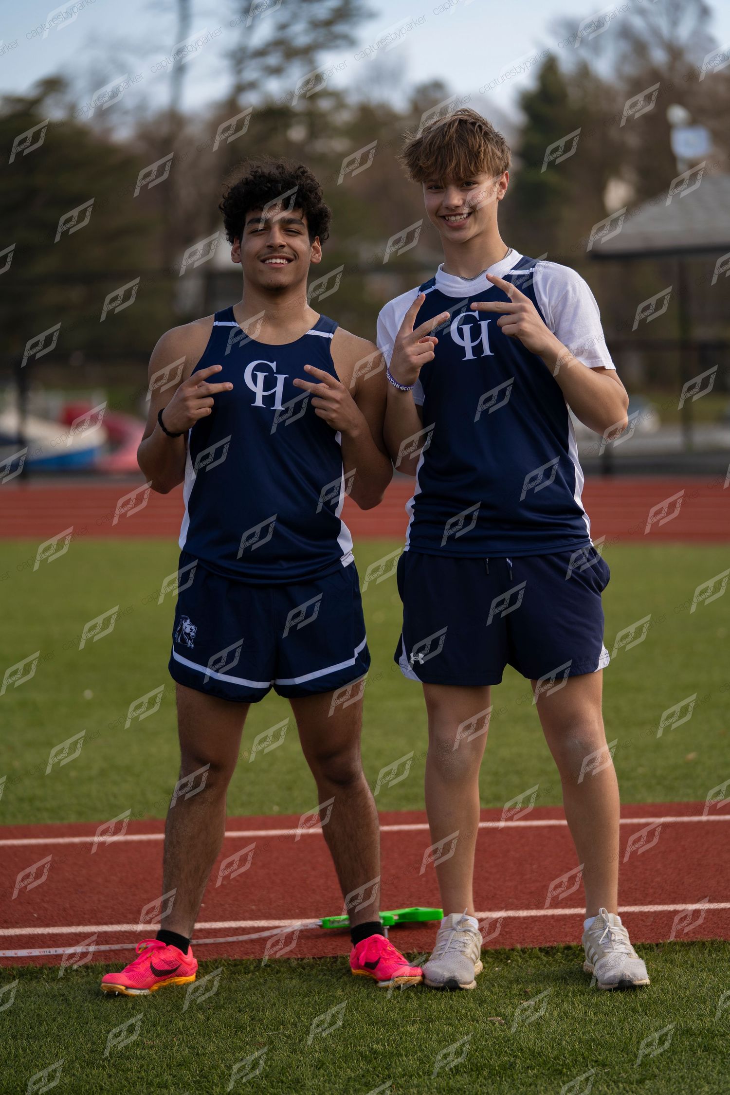 Two male high school track athletes in navy CH uniforms pose together on red track.