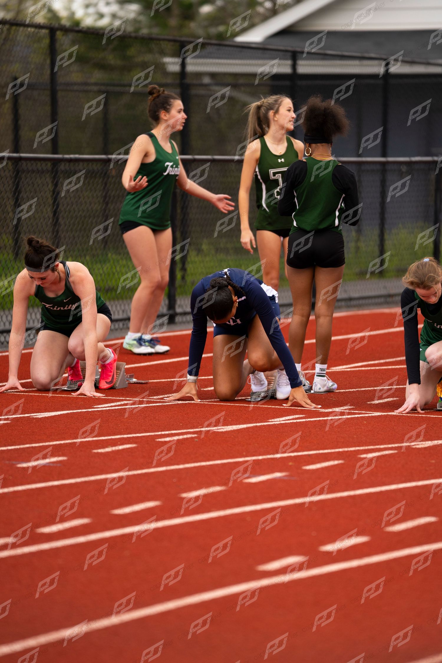 Female athletes prepare at starting blocks on red track during high school track and field meet.