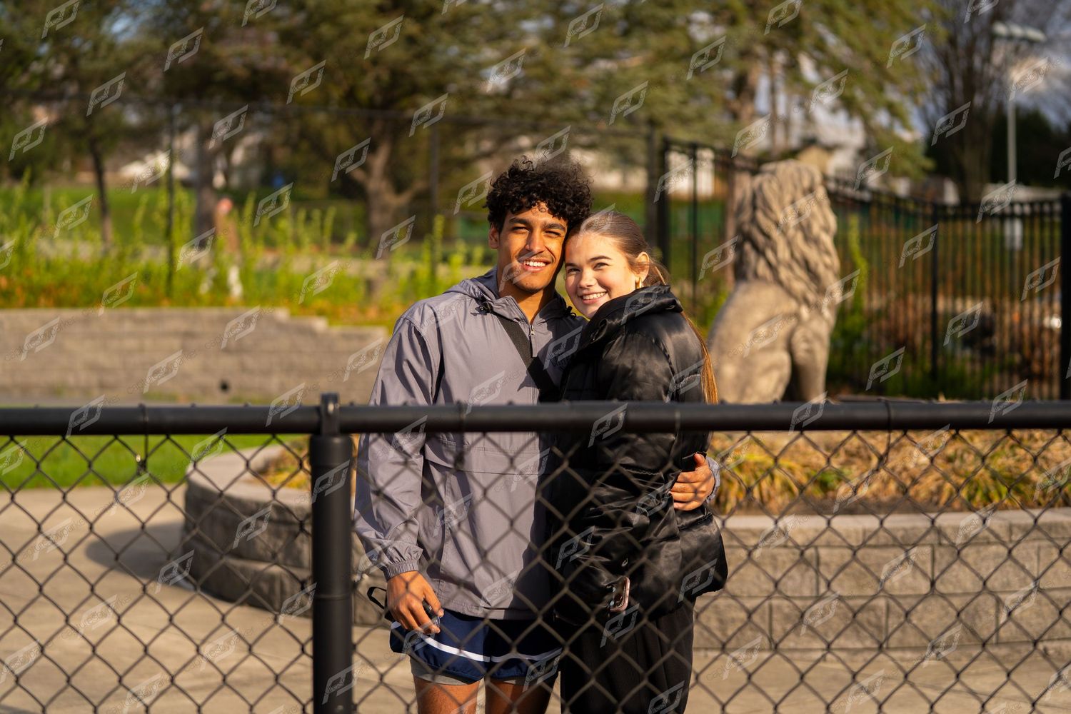Two smiling teenagers posing together near chain-link fence at outdoor track event.