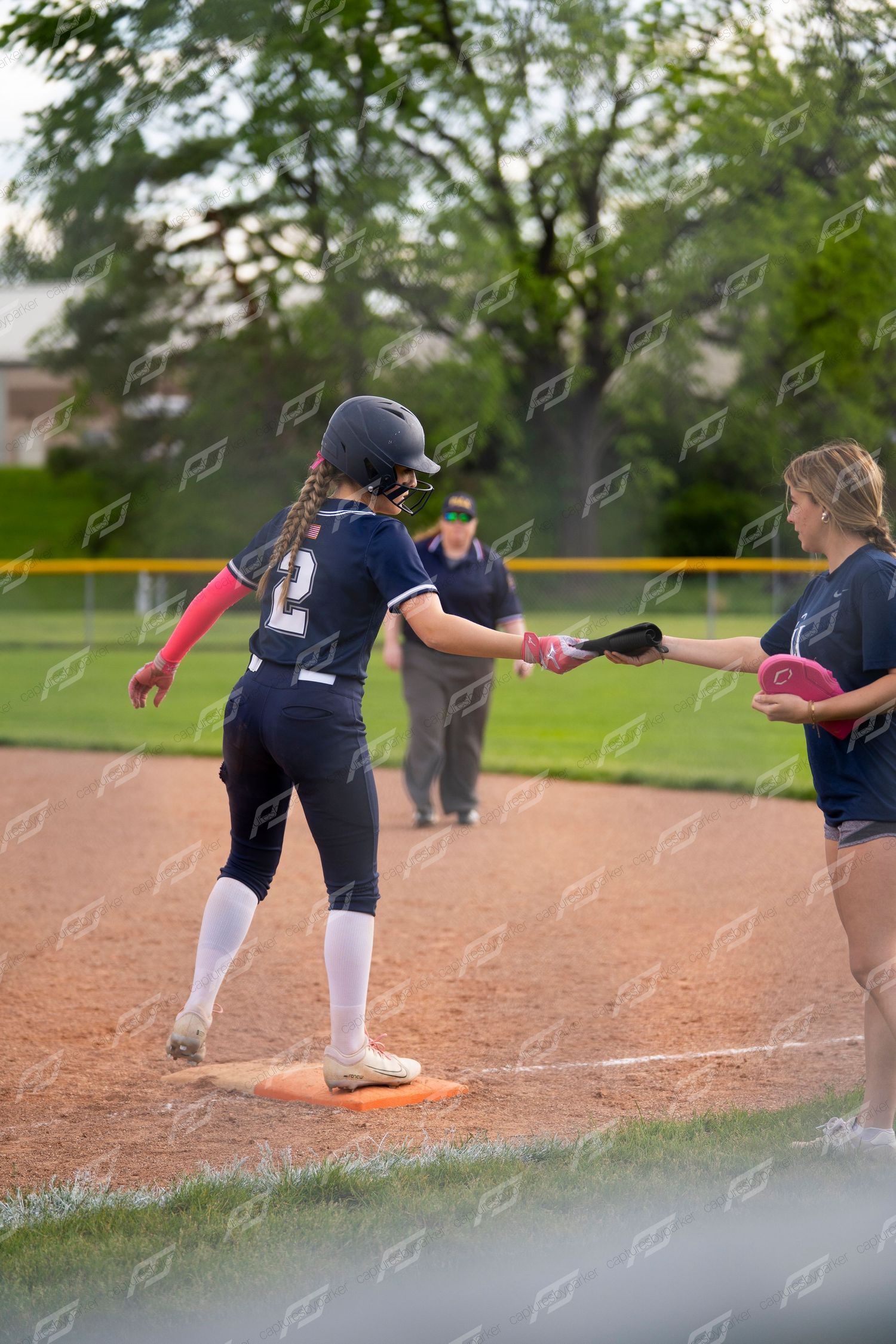 A softball player in navy uniform receives her batting glove from a teammate at first base.