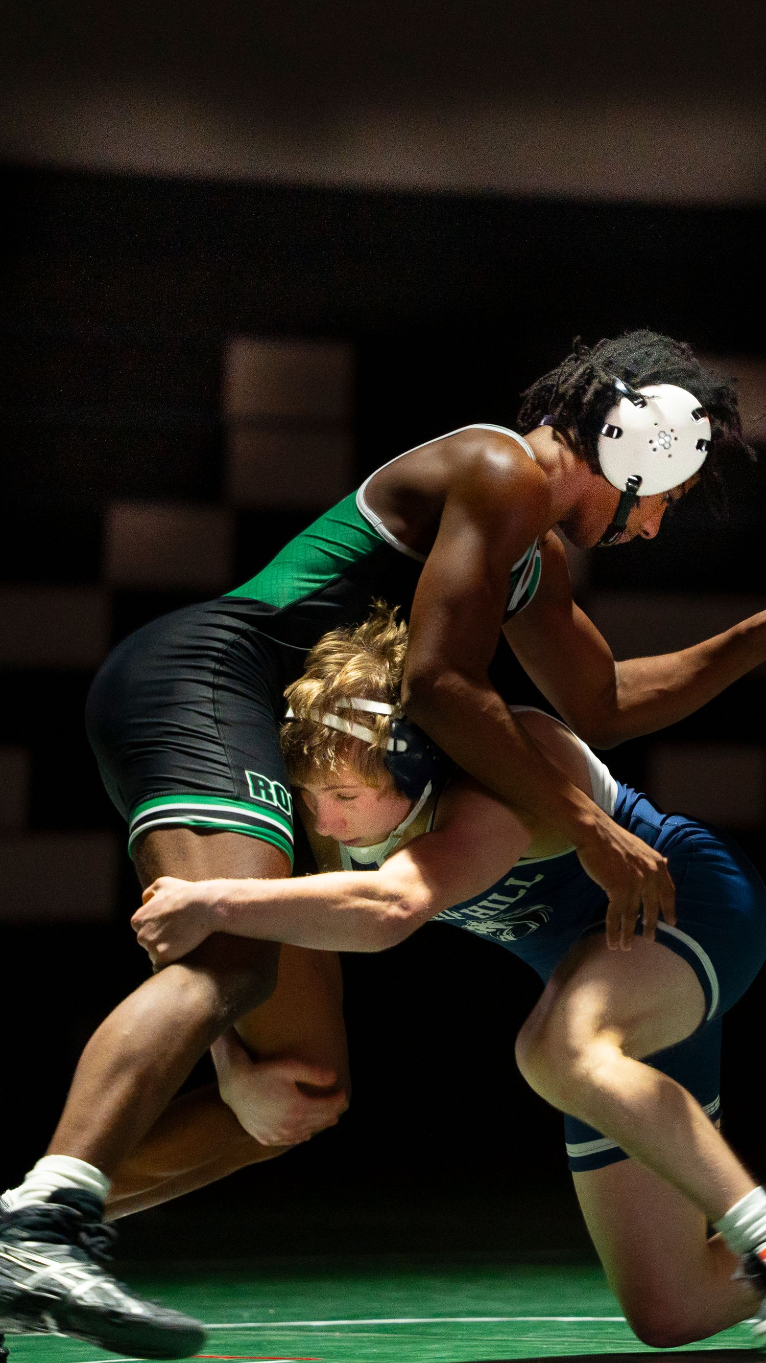 Wrestler in green Trinity singlet grapples with opponent in blue during competitive match.
