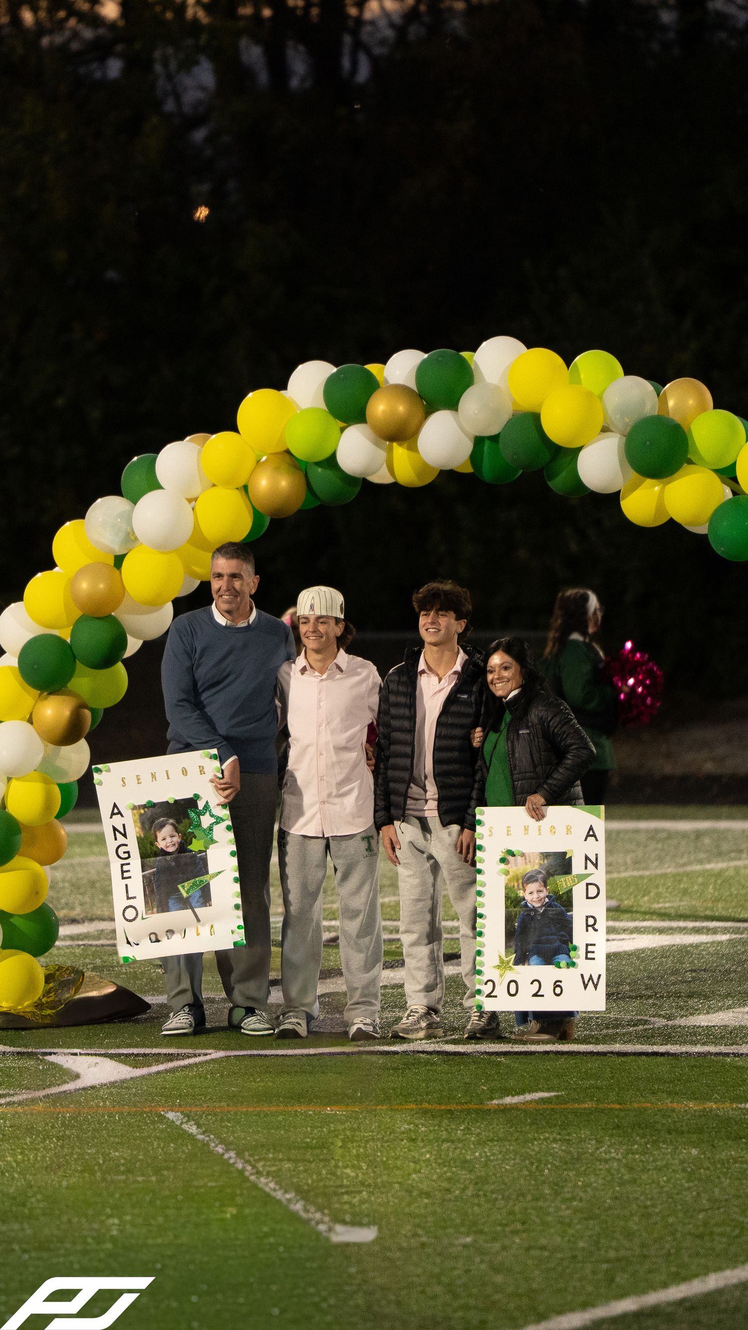 Two seniors named Angelo and Andrew pose with family under a balloon arch at a school senior night event.
