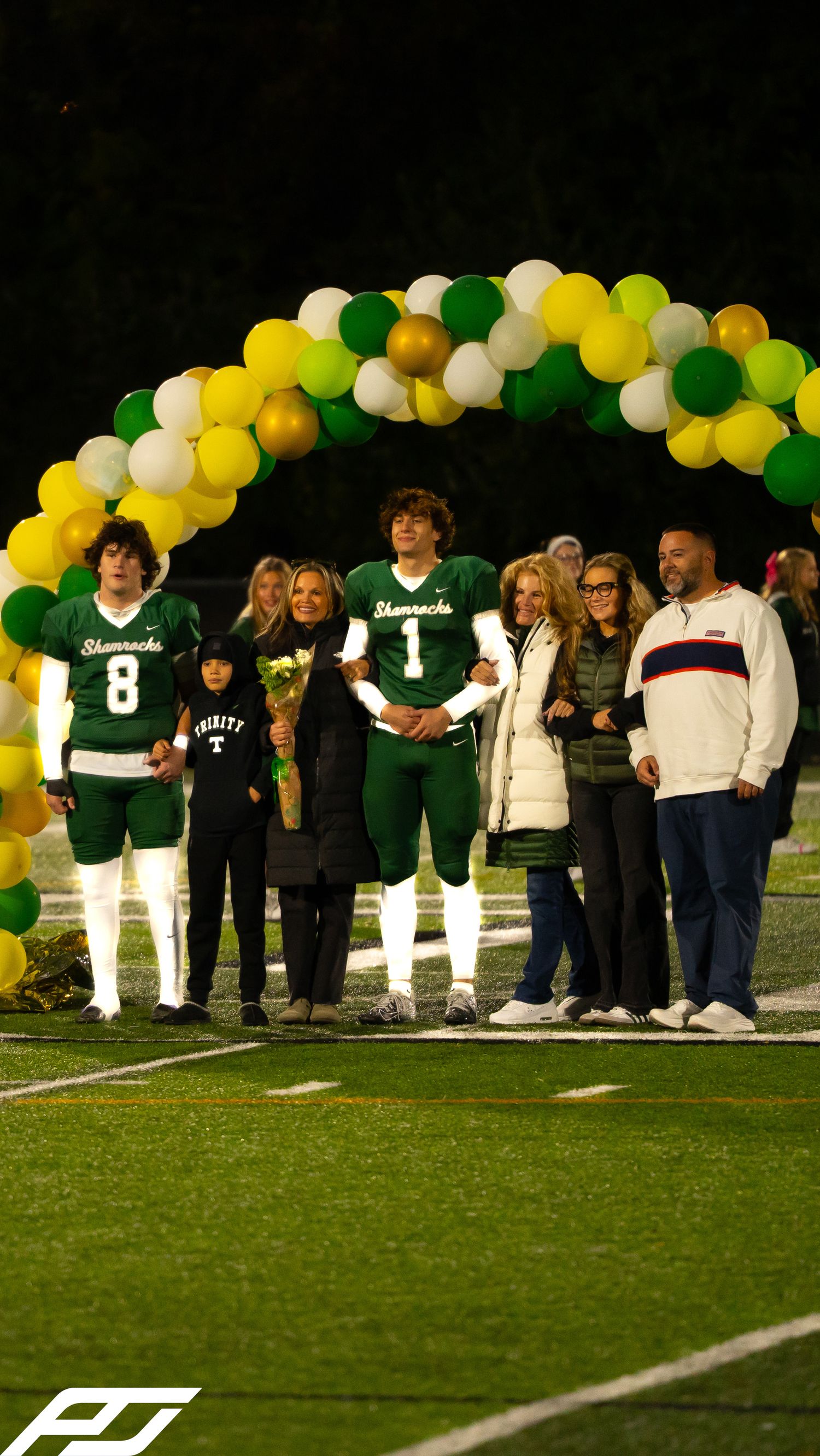 Two Shamrocks football players in green uniforms pose with families under a festive balloon arch at night.