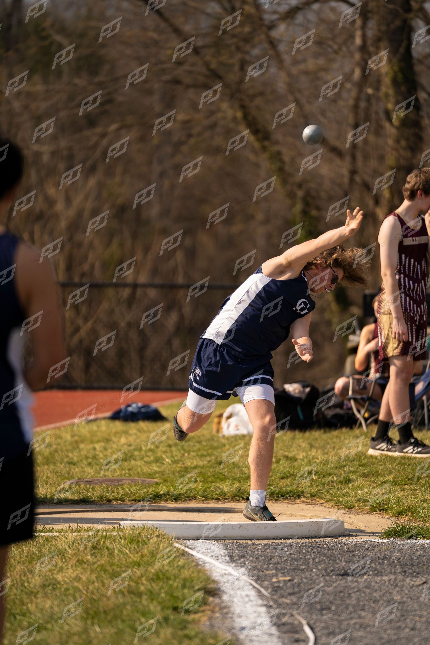 Male athlete in navy uniform throwing shot put at an outdoor high school track meet.