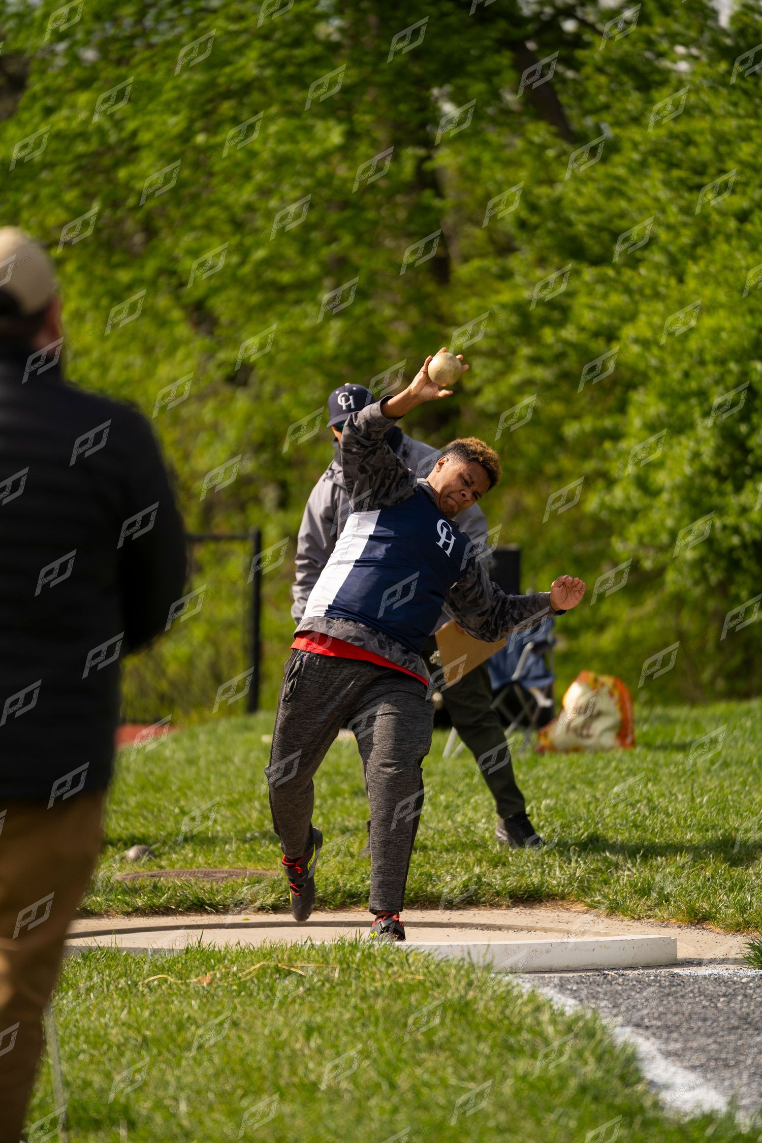Male athlete in navy CH jacket throwing shot put at outdoor track and field competition.