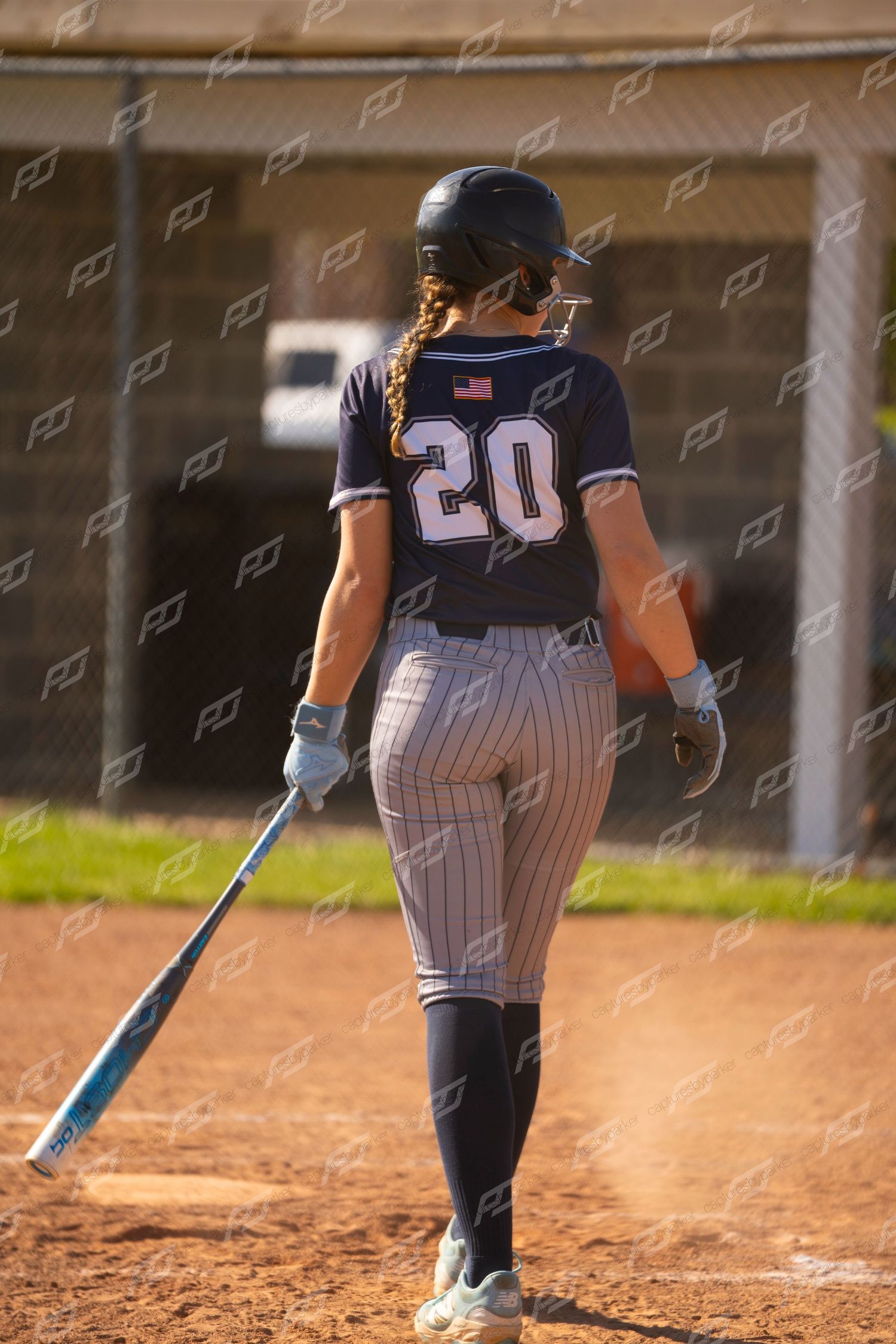 Female softball player #20 in navy pinstripe uniform walking with bat viewed from behind at plate.