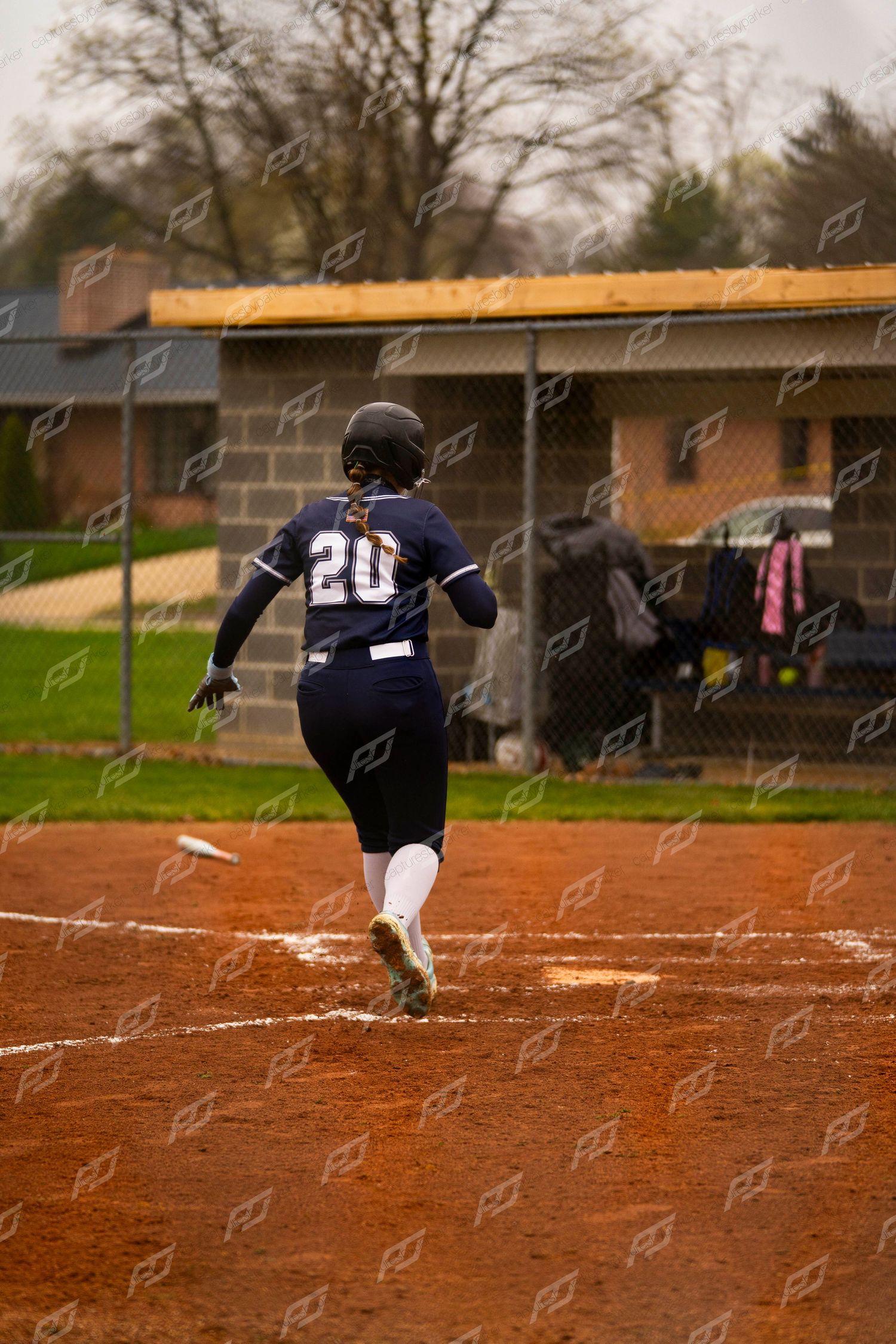 Softball player #20 in navy uniform sprints down baseline after hitting ball during game.
