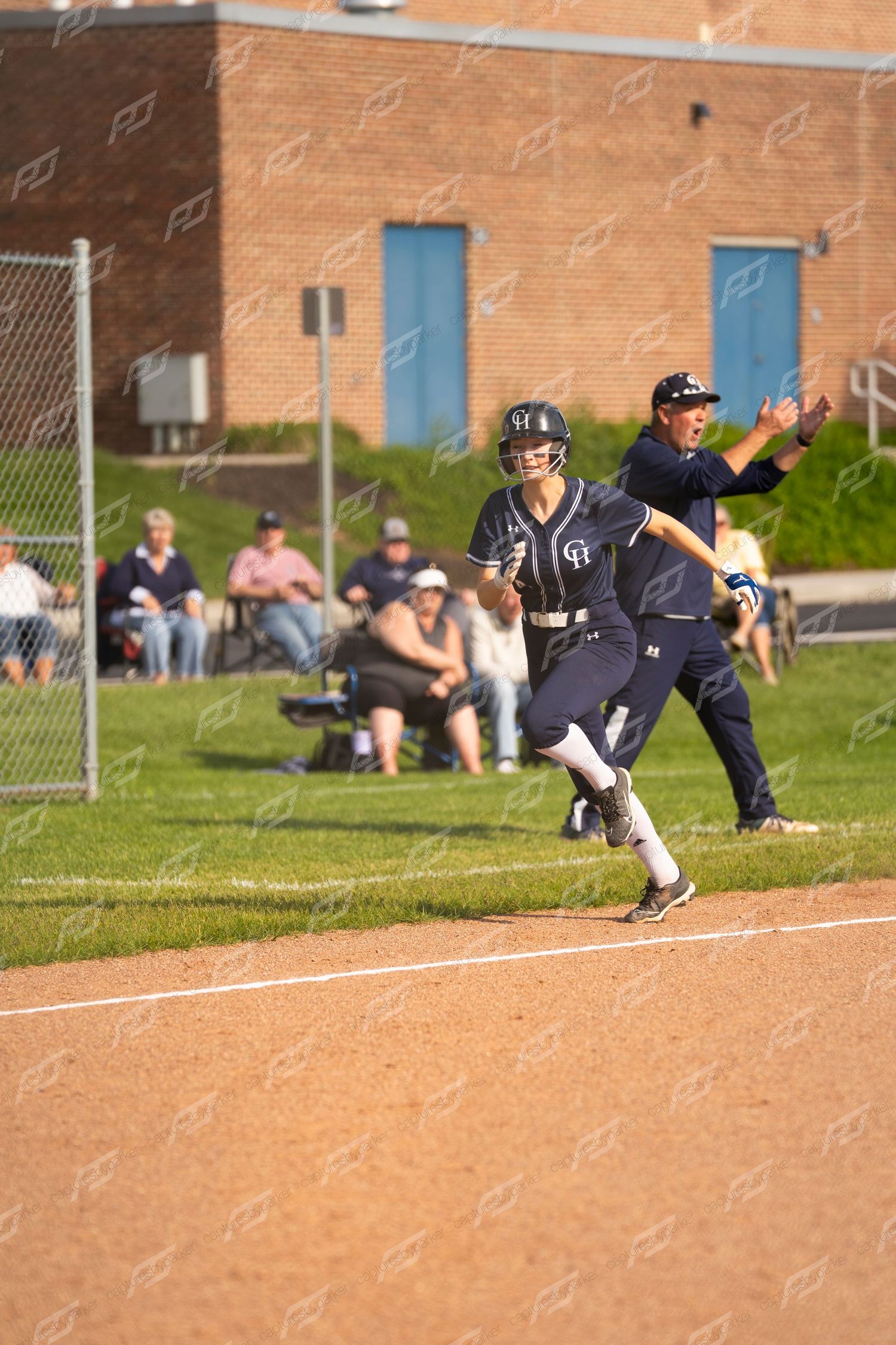 A softball player in navy uniform runs the bases as a coach signals during a competitive game.