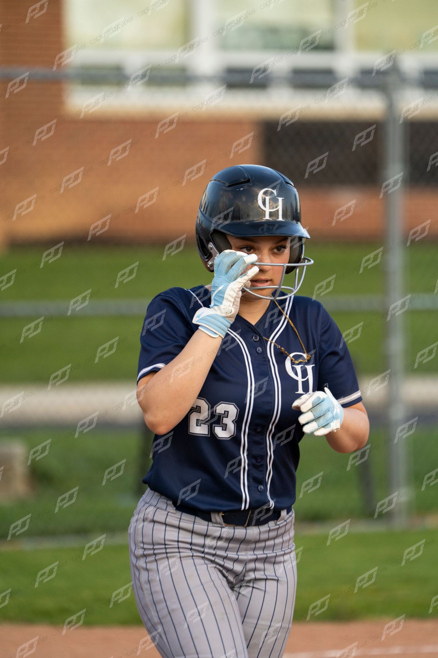 Female softball player number 23 in navy CH uniform adjusts helmet face guard on field.