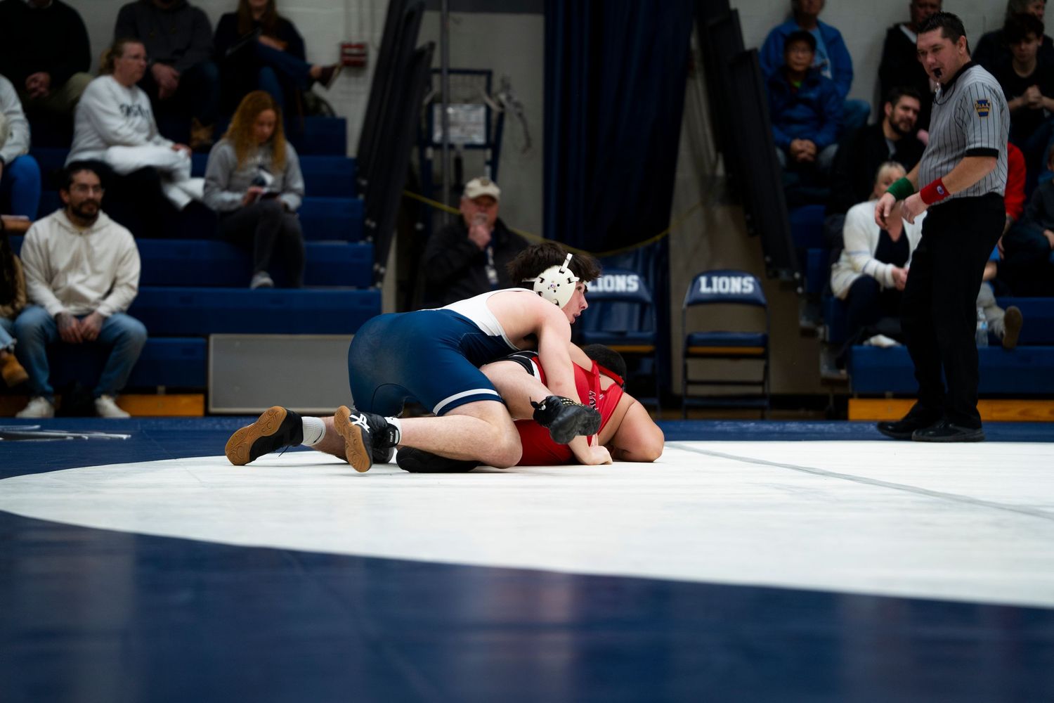 High school wrestler in blue singlet pins red-uniformed opponent to mat as referee watches closely.