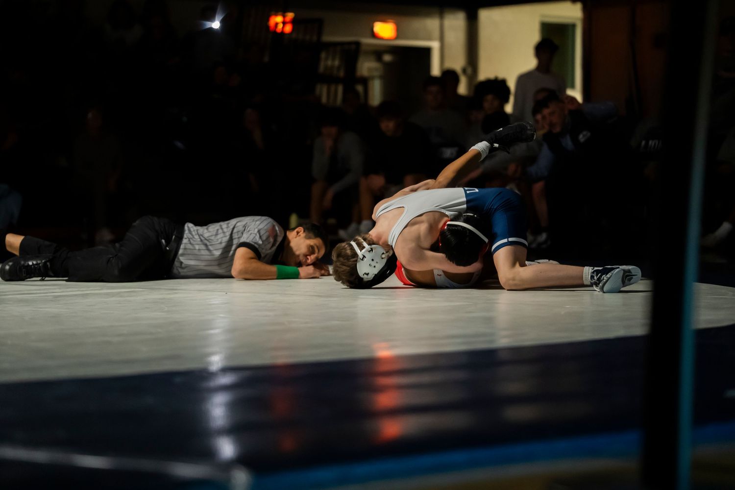 Two wrestlers lie exhausted on the mat after an intense wrestling match in a darkened gymnasium.