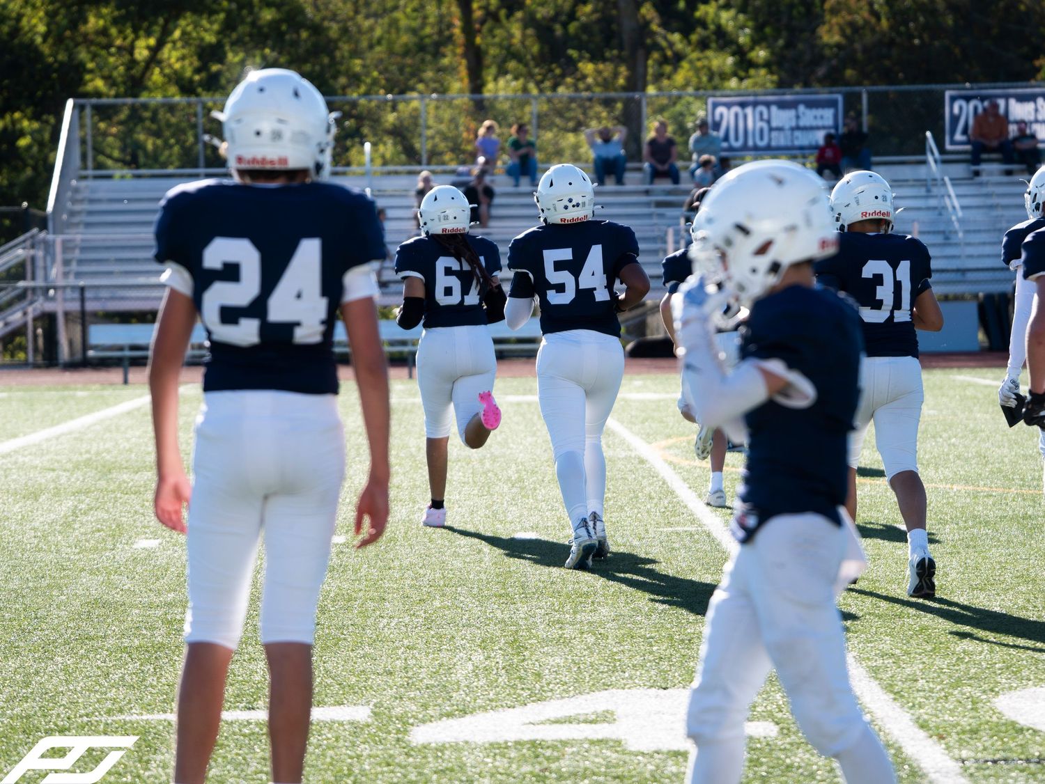 Youth football players in navy and white uniforms walk across green turf field during a game day.