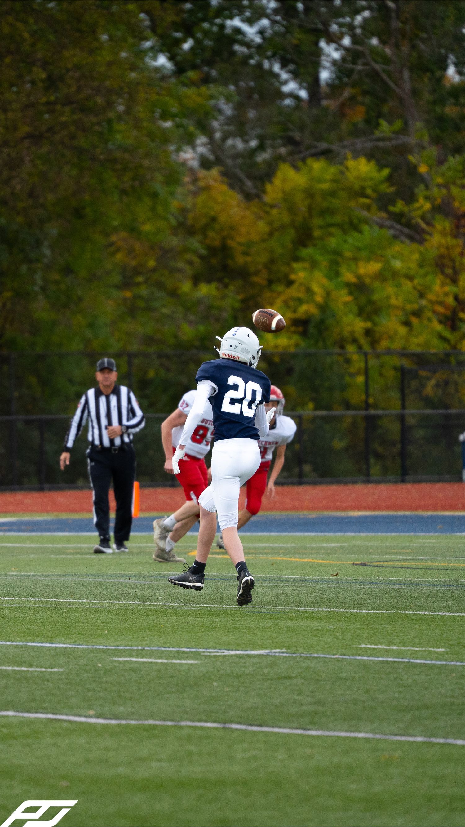 Youth football player number 20 in navy uniform catches a pass on a green turf field with referee nearby.
