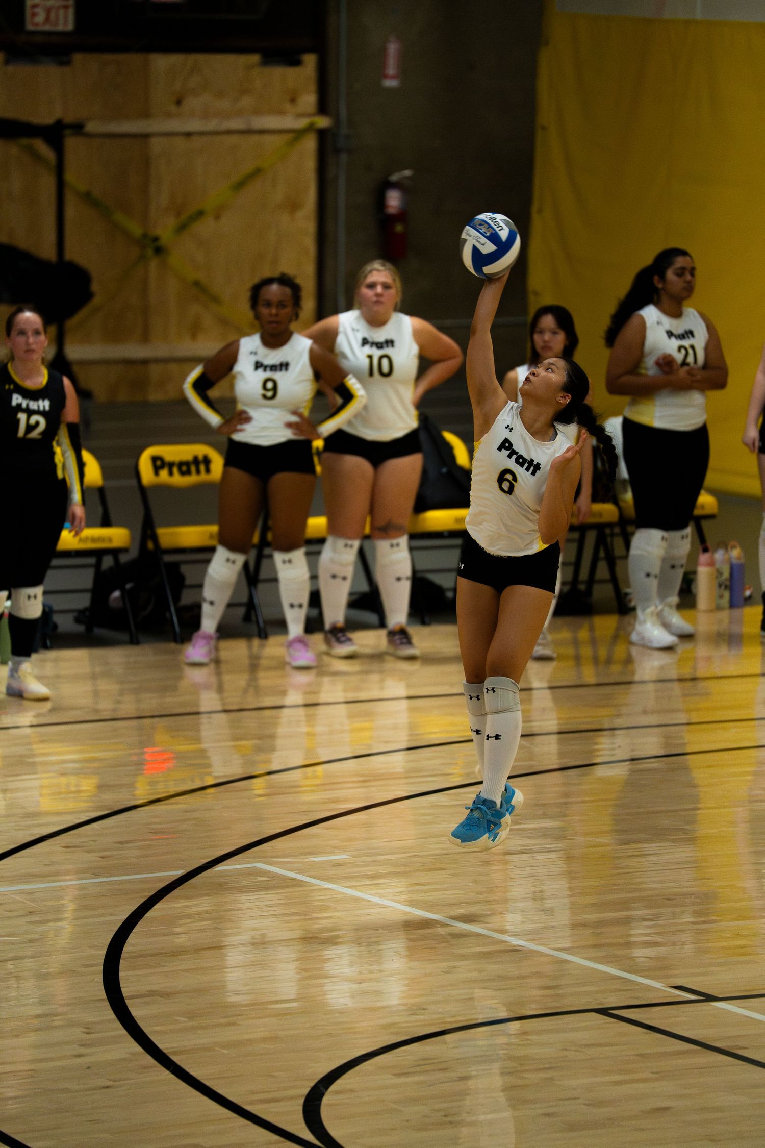 Pratt volleyball player #6 serves the ball during a match, teammates in white and black uniforms visible on the court.