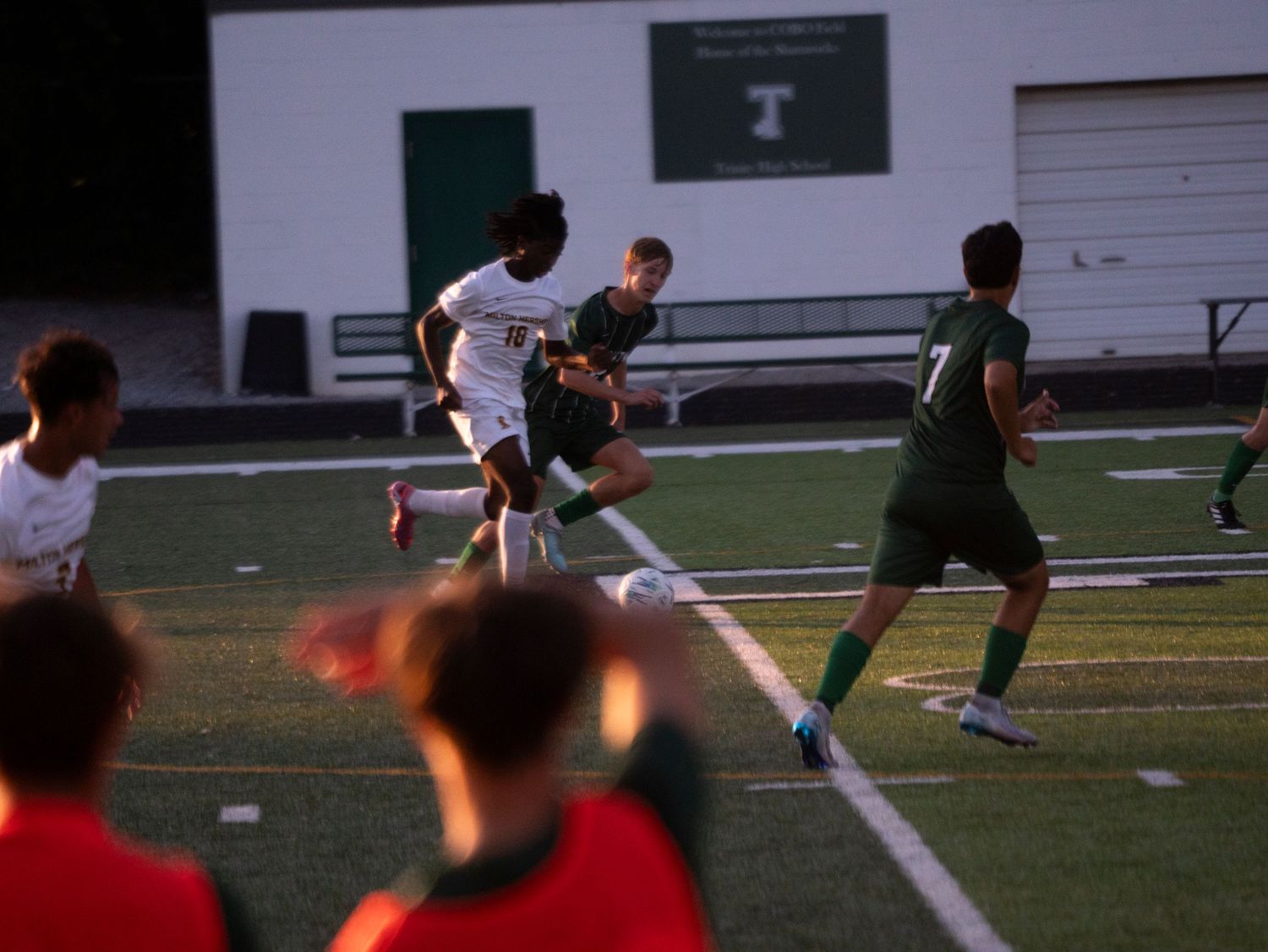 High school soccer players in green and white uniforms battle for possession near the goal area.