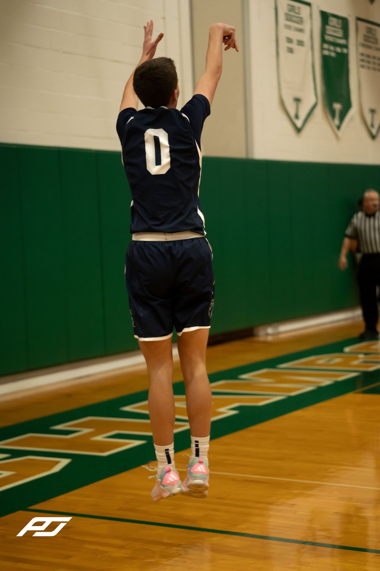 Player number 0 in navy uniform jumps to shoot a basketball in a green gymnasium.