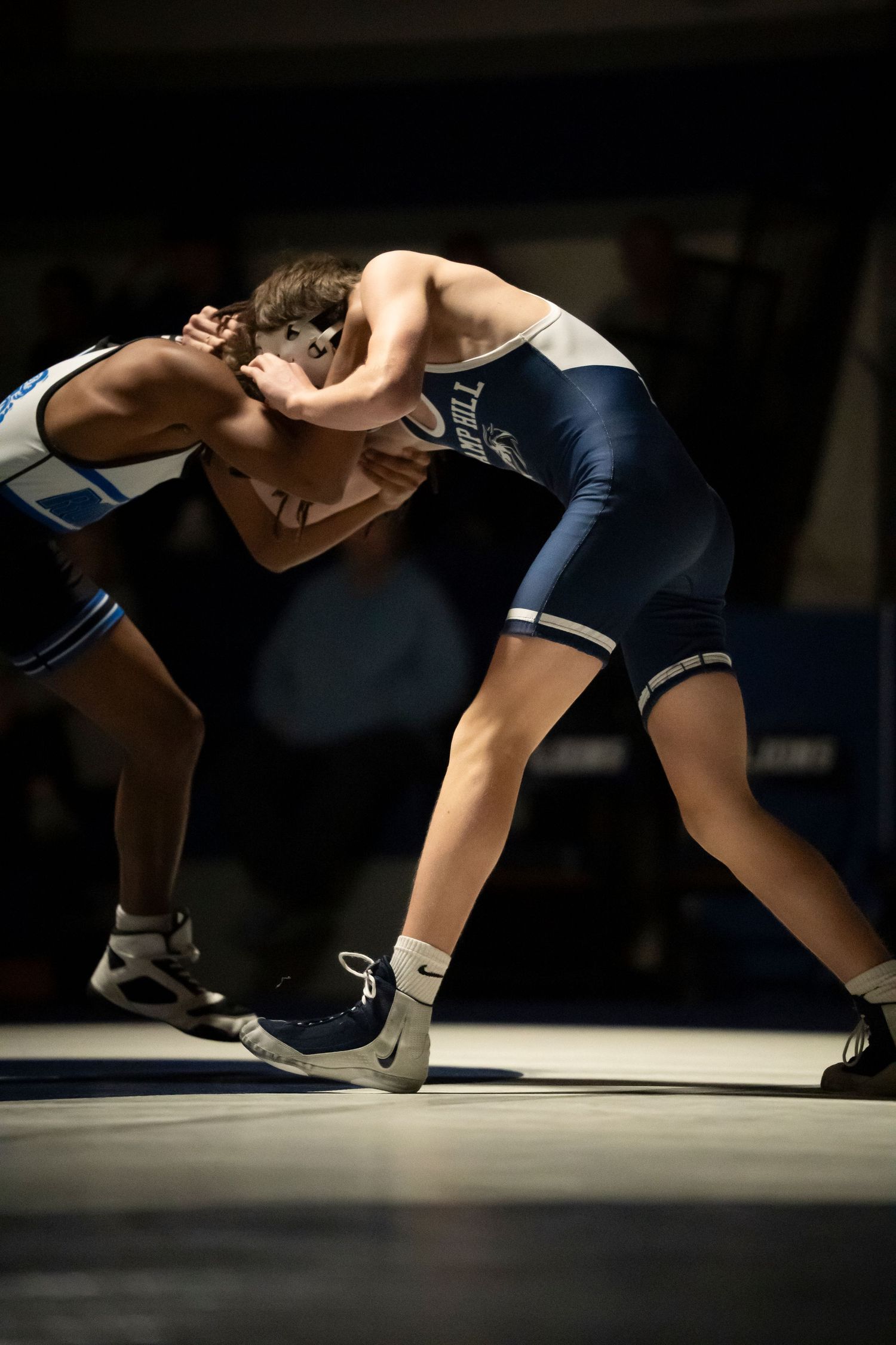 Two wrestlers battle in a tie-up clinch position during a high school wrestling match under dramatic lighting.