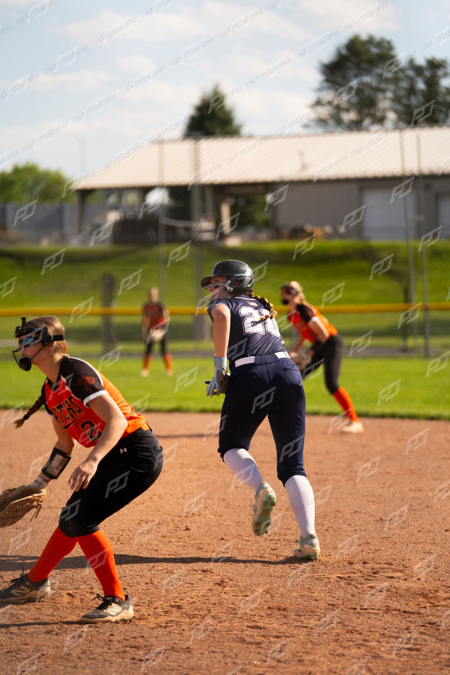 Navy-uniformed softball player rounds base past orange-uniformed fielder during outdoor game.