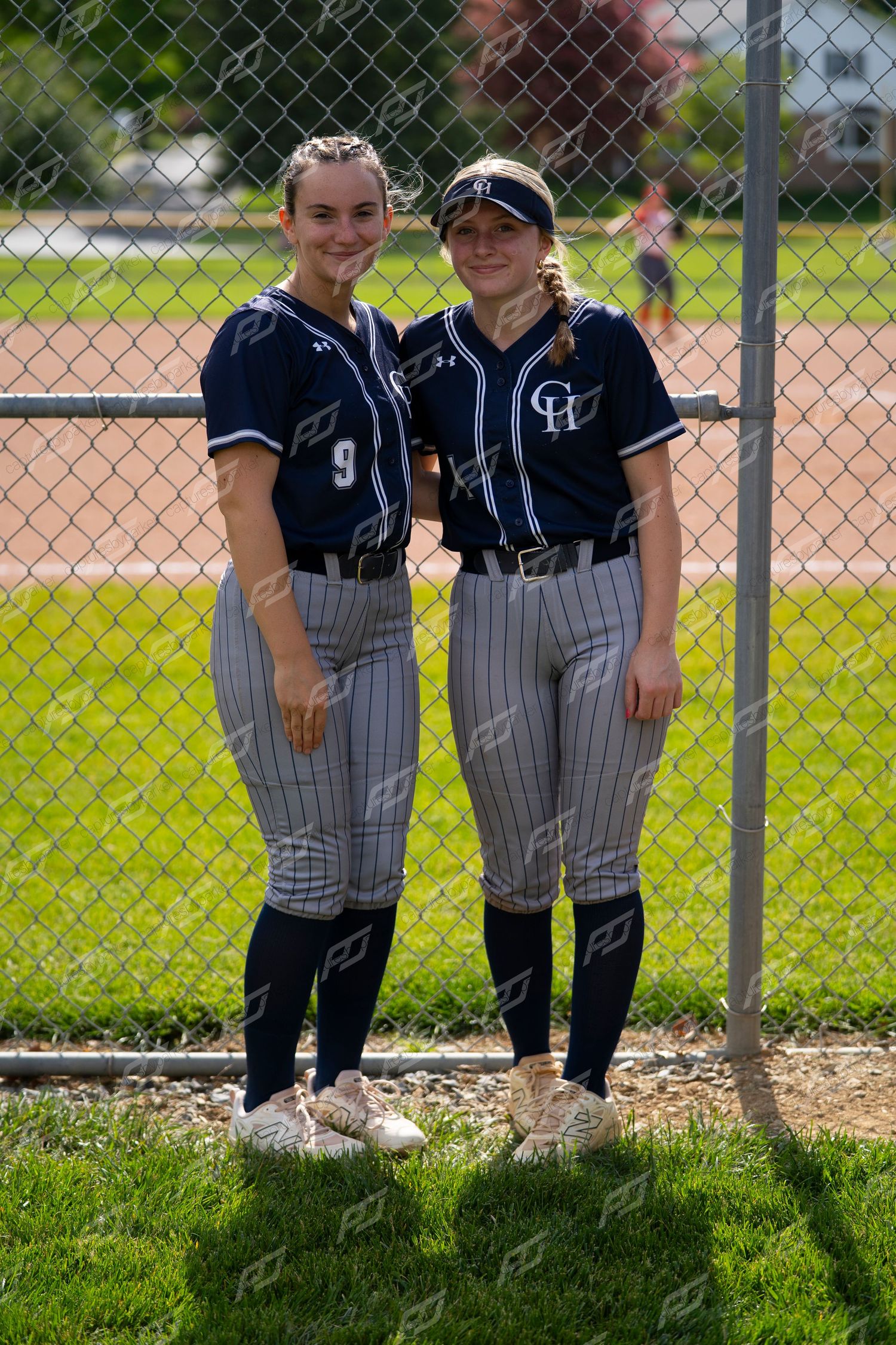 Two female softball players in navy GH uniforms pose together by a chain-link fence.