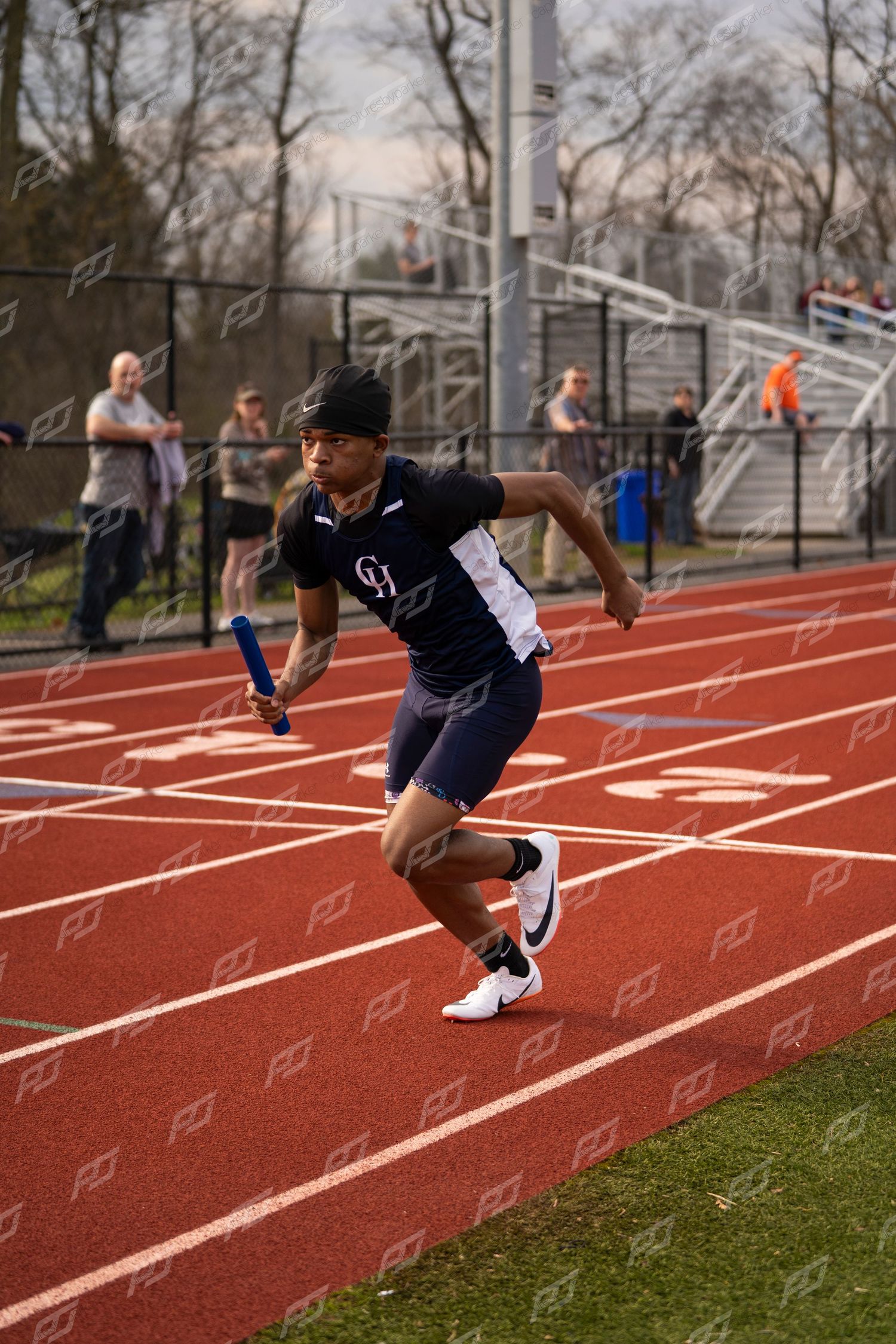 Male sprinter races on red track holding blue relay baton during outdoor track meet.