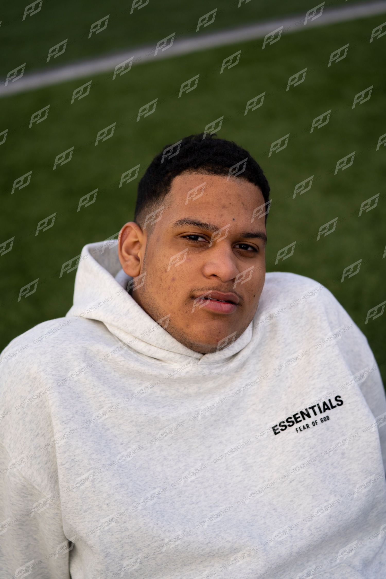 Young man wearing a white Fear of God Essentials hoodie poses in front of a green turf field.