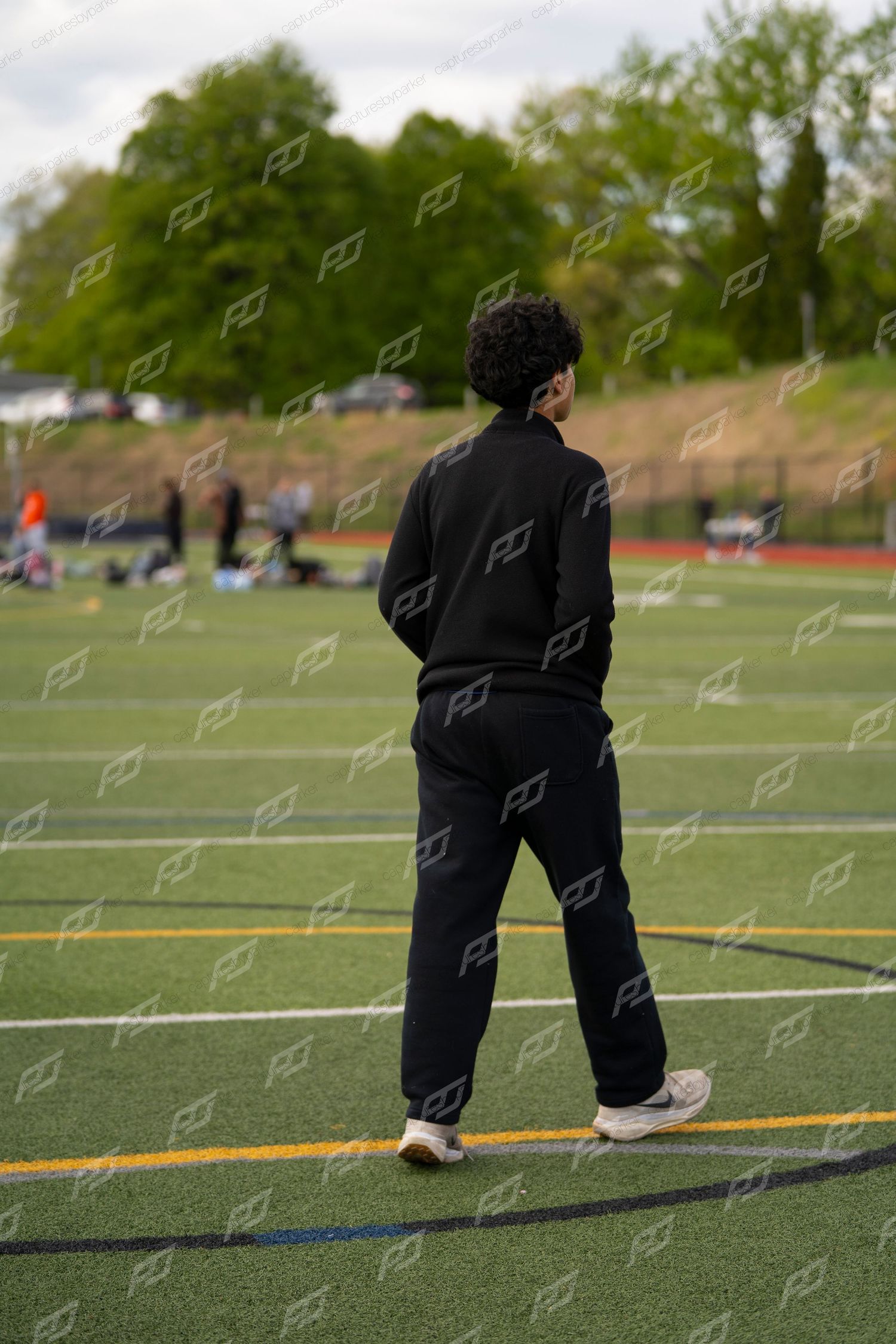 Athlete in black tracksuit standing on green turf field viewed from behind at outdoor sports event.