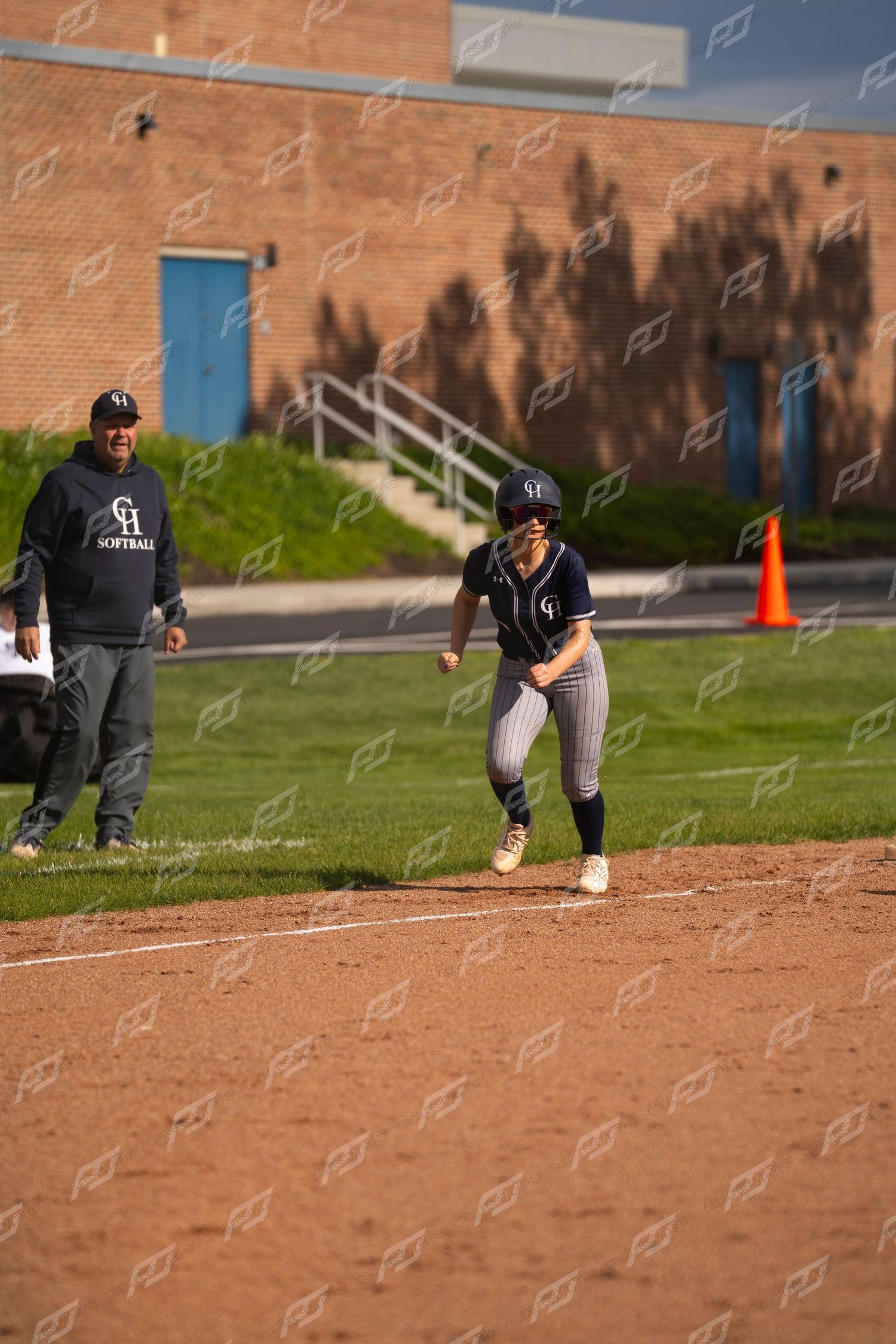 Female softball player leads off base while coach watches during a high school softball game.