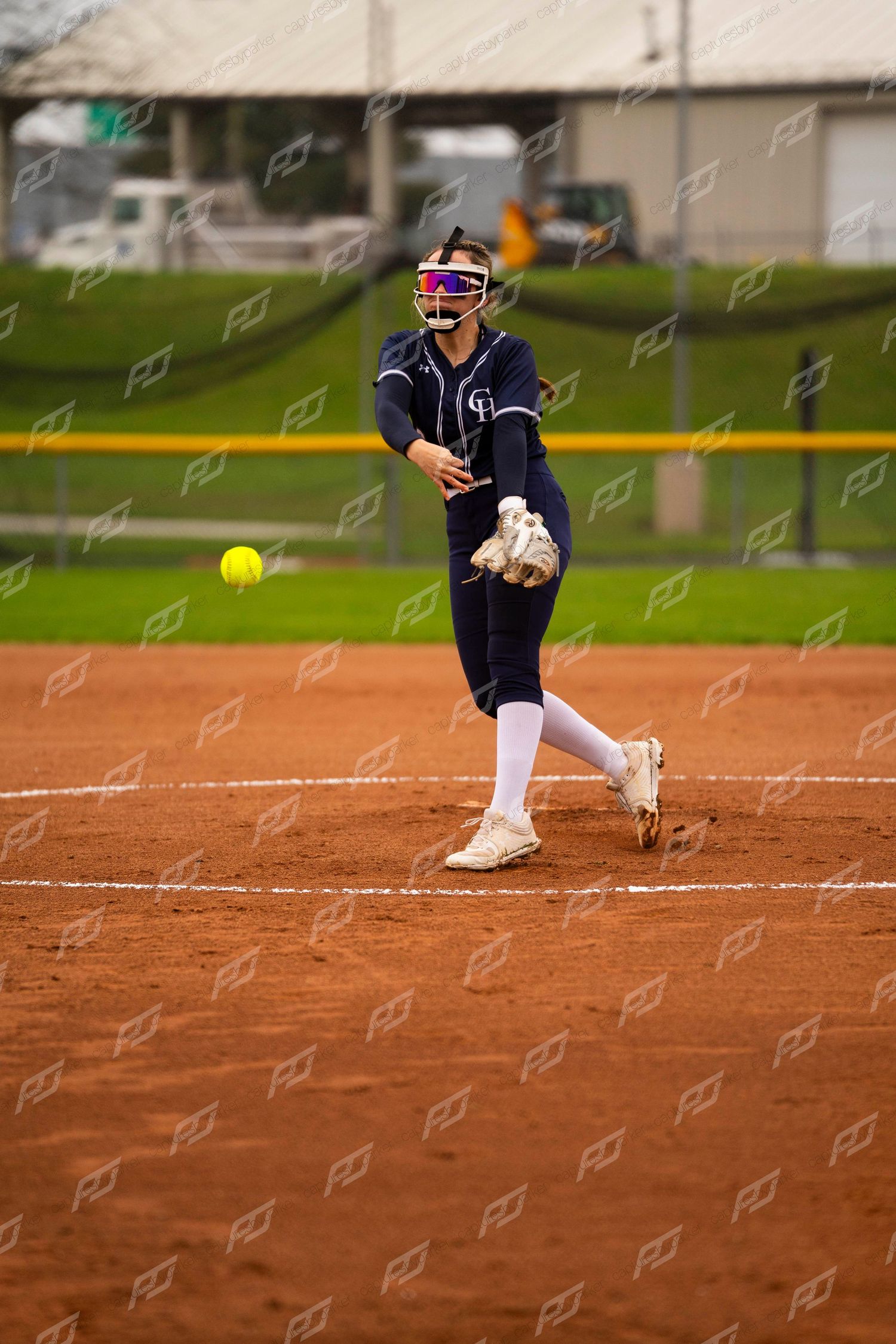 Softball pitcher in navy uniform releasing a yellow ball during a windmill pitch on a red clay field.