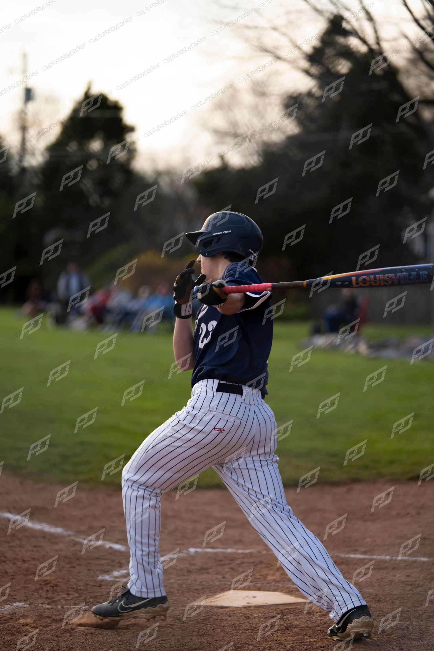 Baseball player in white pinstripe uniform swings a Louisville Slugger bat during a game at dusk.