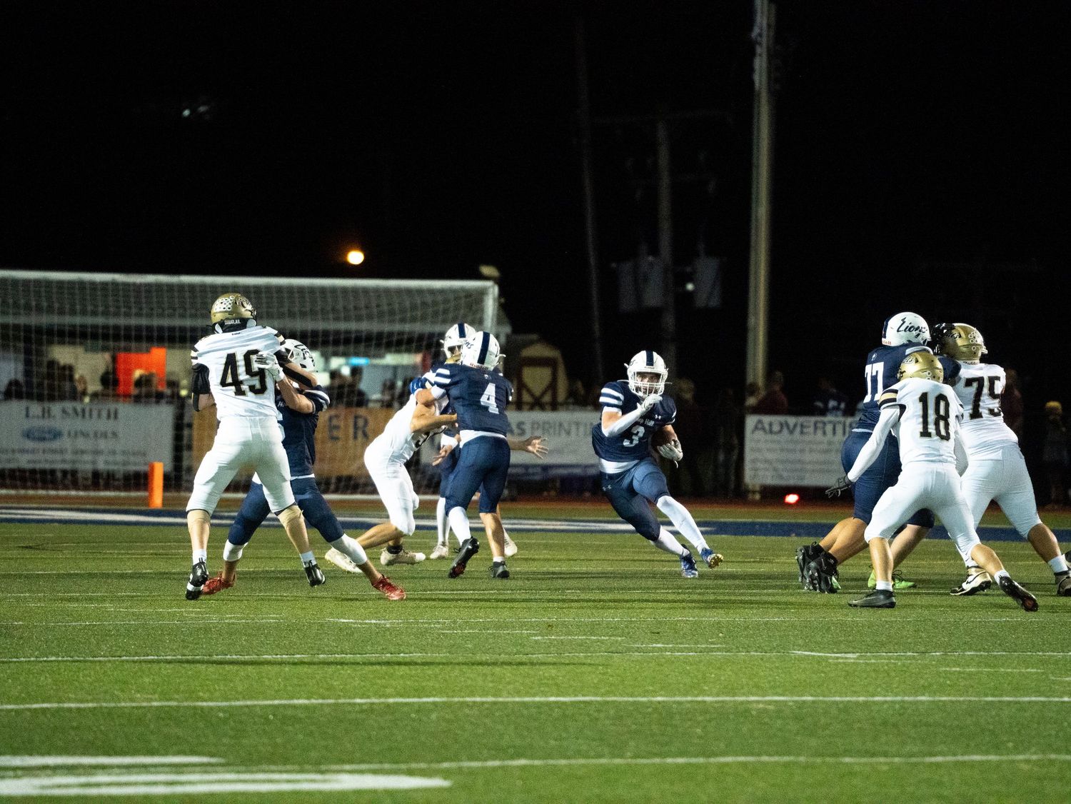 Night high school football game action shot with players in navy and white uniforms competing on a lit field.