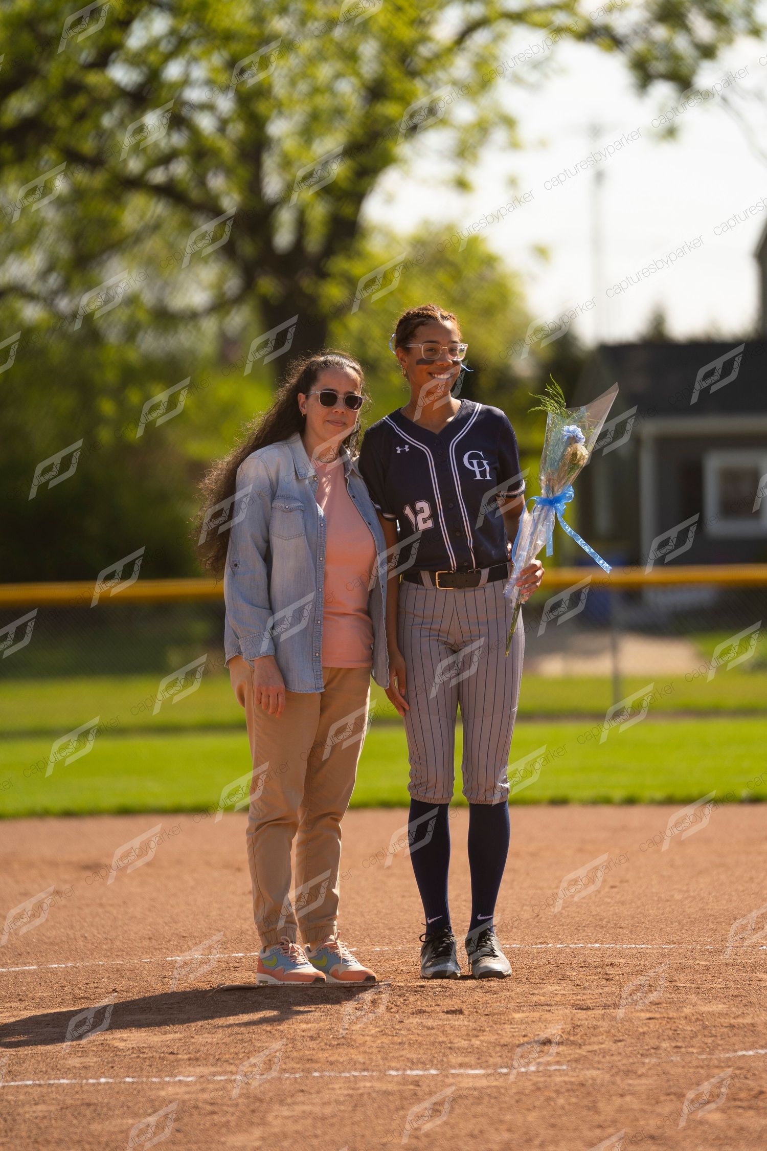 Softball player in navy uniform number 12 holds flowers while posing with parent on sunny field.