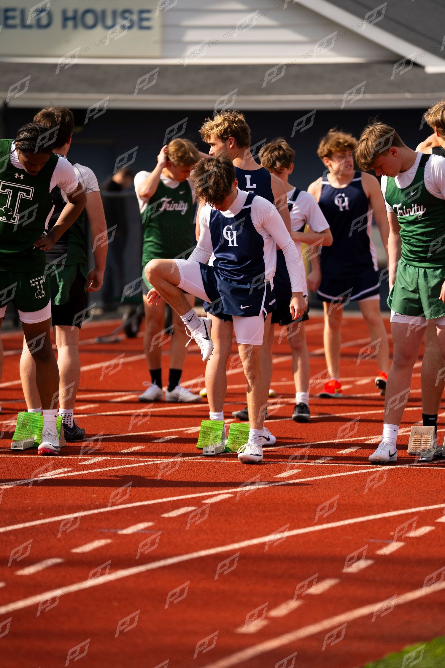 Male track athletes in green and navy uniforms warm up at starting blocks on red outdoor track.
