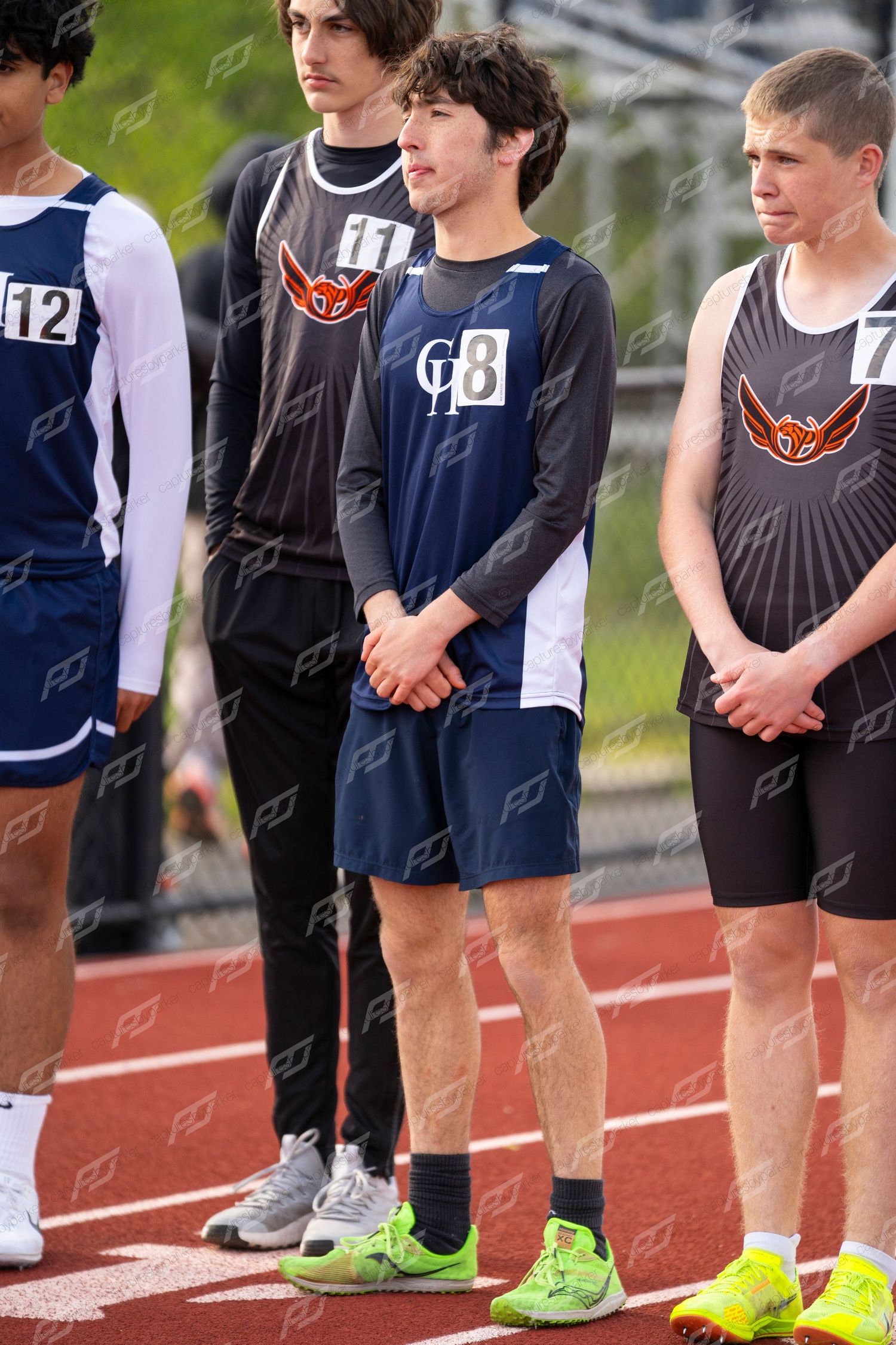 Young male track athletes stand on a red track before a race wearing numbered bibs.