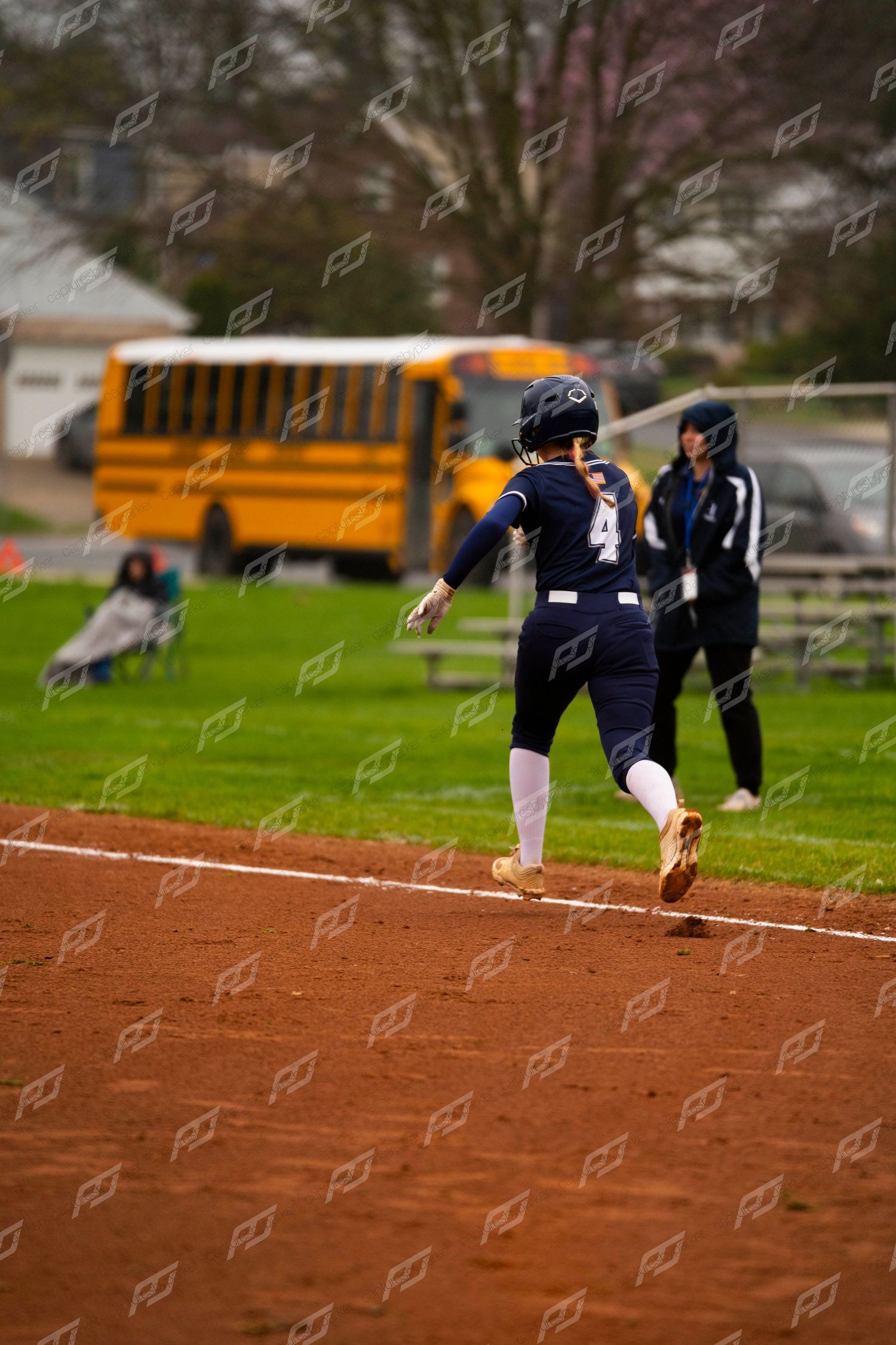 Female softball player #4 in navy uniform runs the bases with a yellow school bus visible in background.