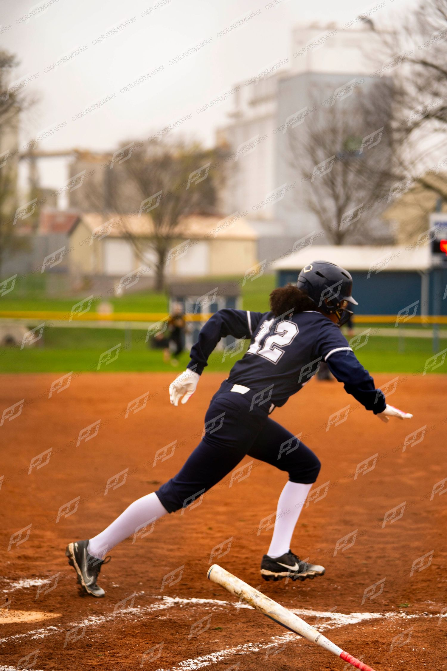 Softball player number 12 in navy uniform sprints from home plate after hitting, bat on ground behind her.