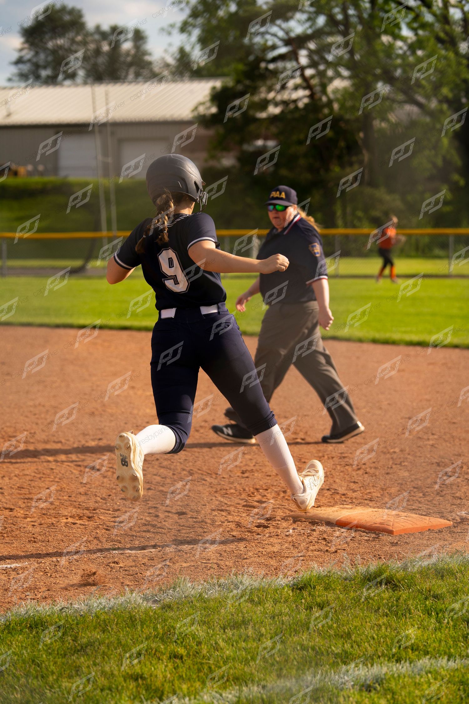 Softball player #9 rounds first base as umpire signals during a game.