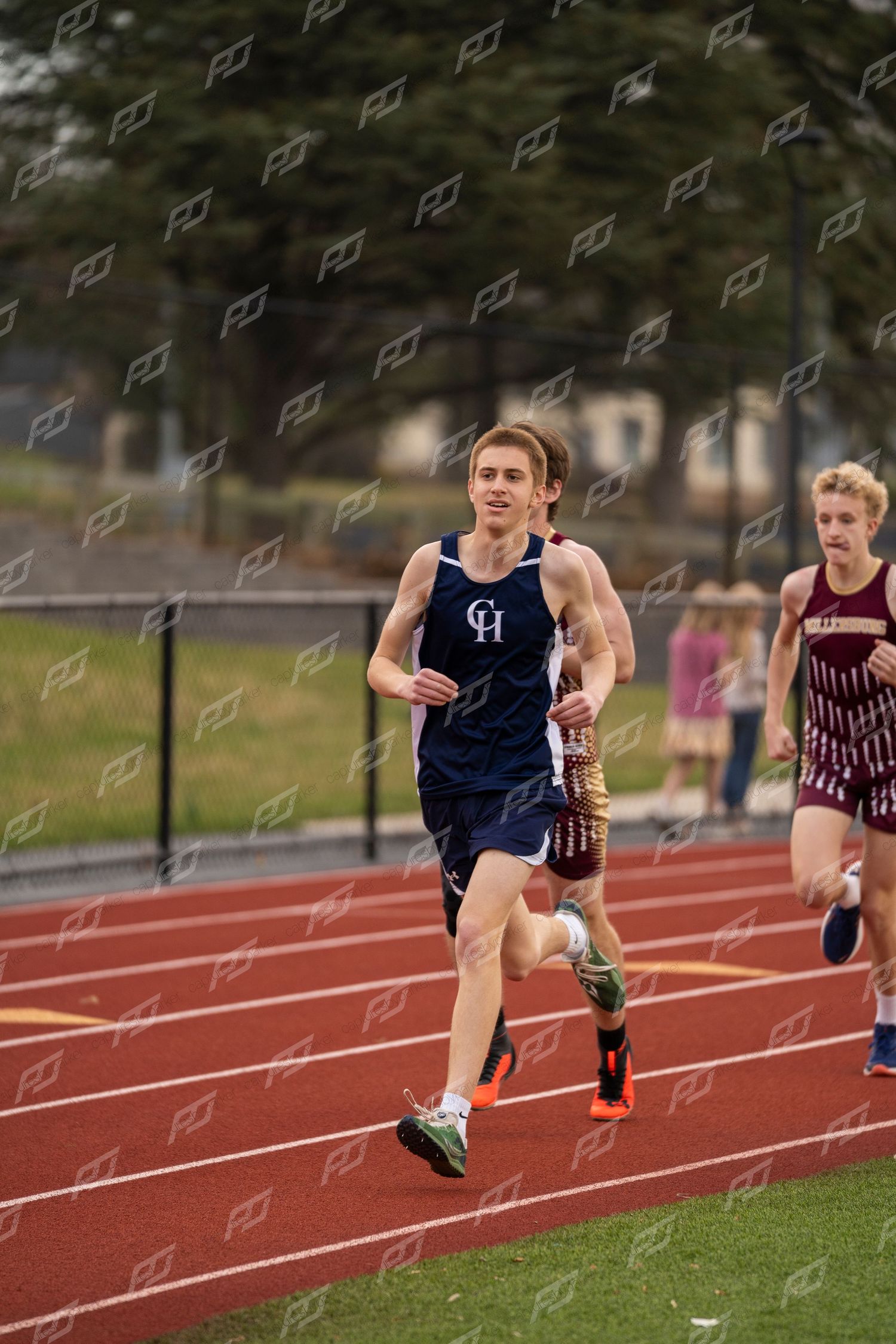 A high school runner in a blue CH uniform leads a race on a red outdoor track.
