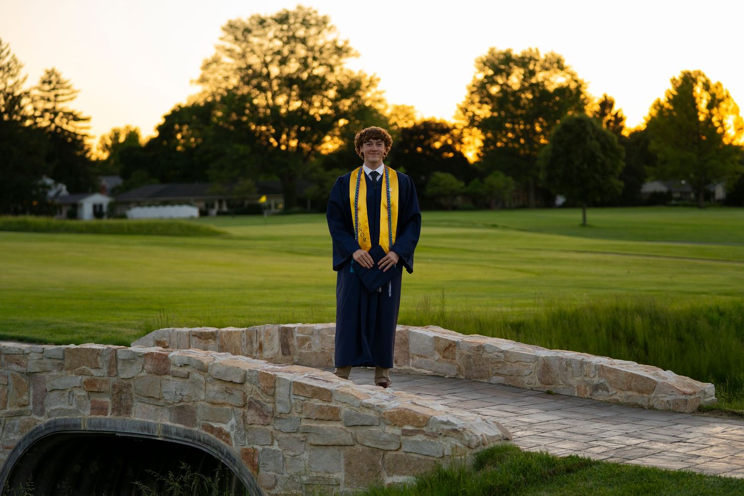 Graduate in navy blue cap and gown with gold stole stands on stone bridge at golden hour.