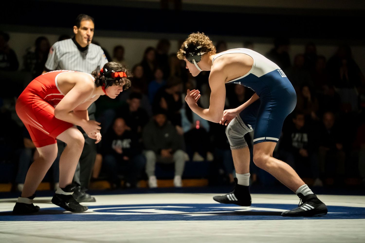 Two wrestlers in red and blue singlets face each other in a ready stance during a match.